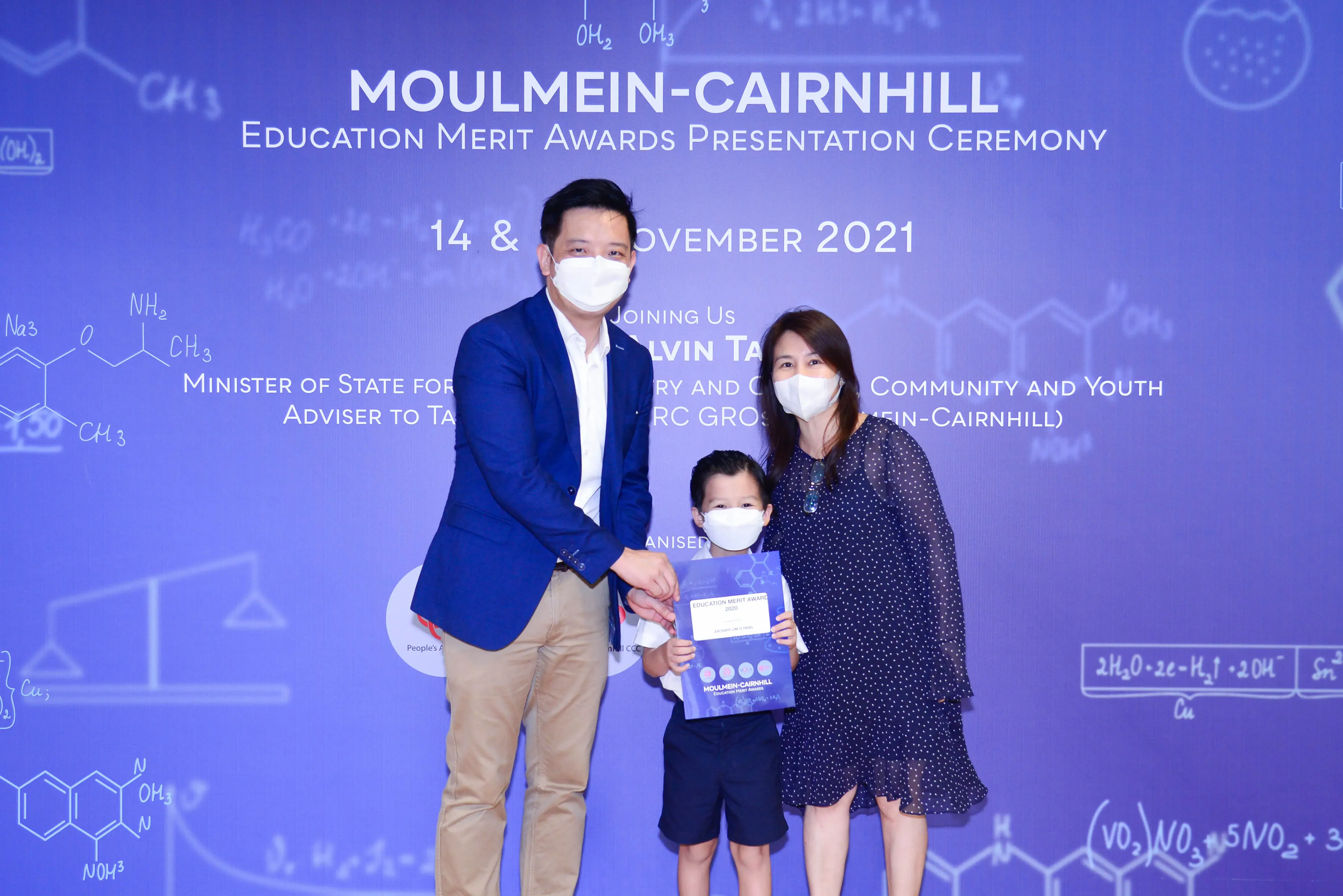 Two masked adults and a masked child holding an award certificate at the Moulmein-Cairnhill Education Merit Awards 2021.