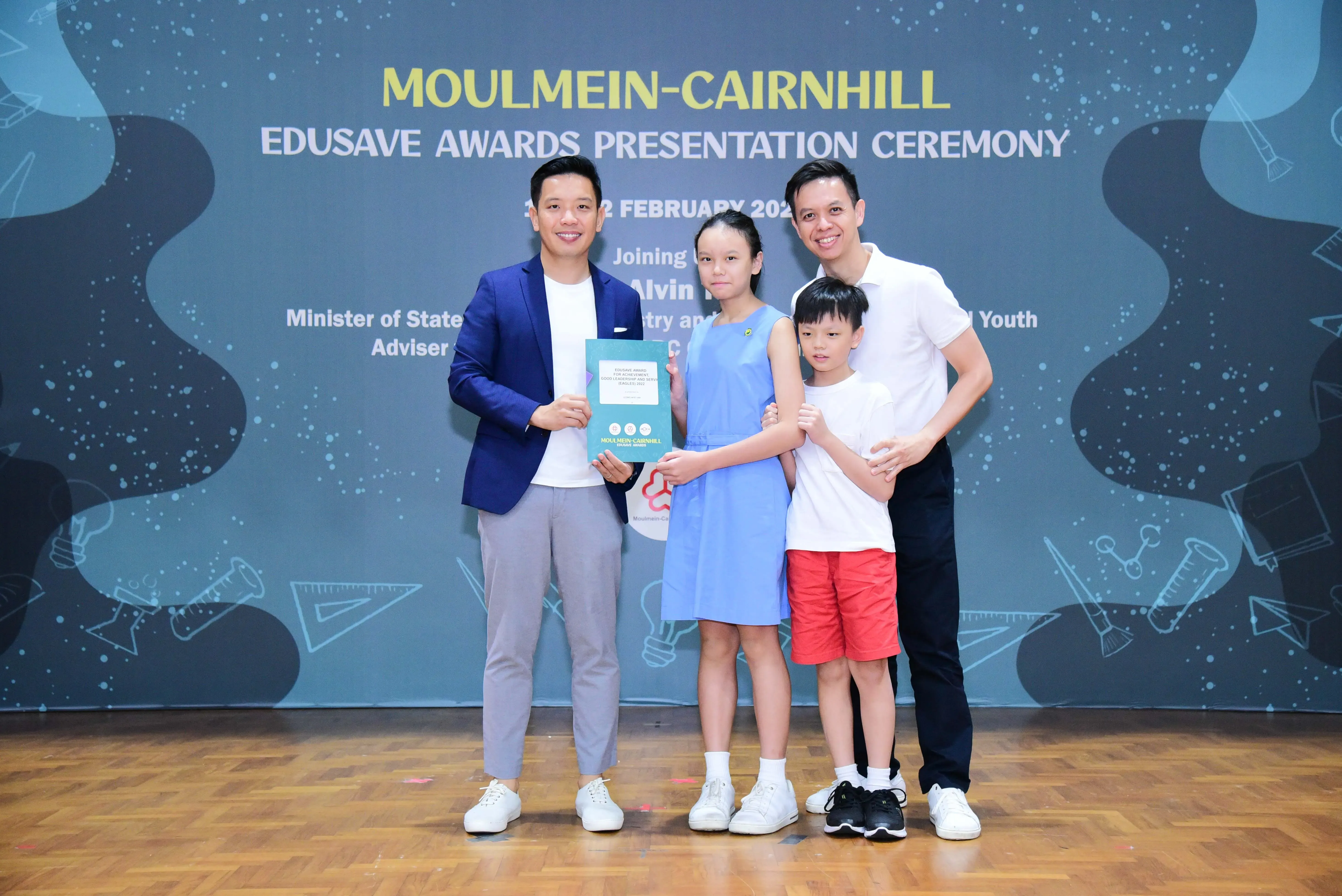 Four people pose at the Moulmein-Cairnhill Edusave Awards Presentation Ceremony, with a girl holding a certificate.