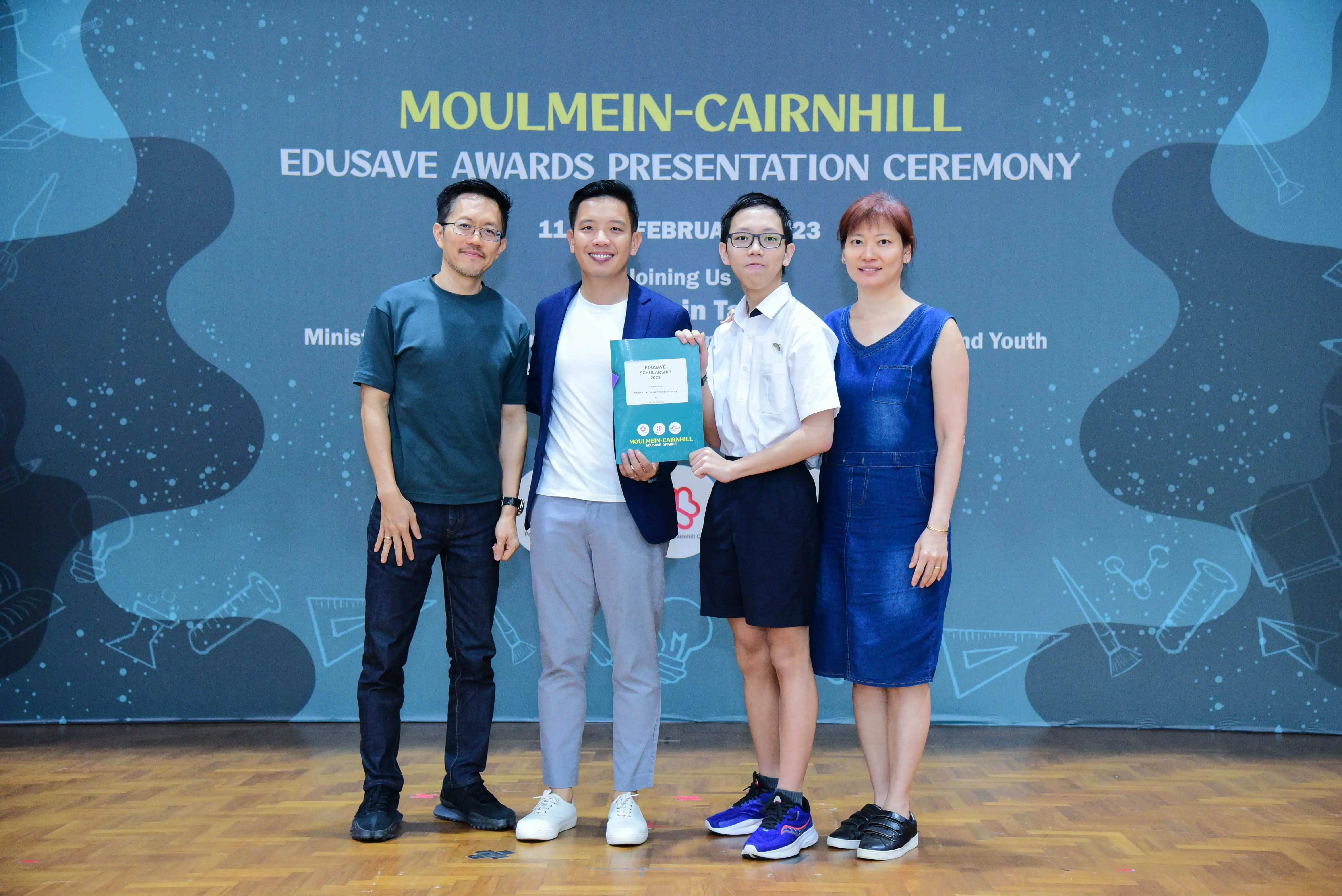 Four people pose holding an award booklet at the Moulmein-Cairnhill Edusave Awards Presentation Ceremony backdrop.