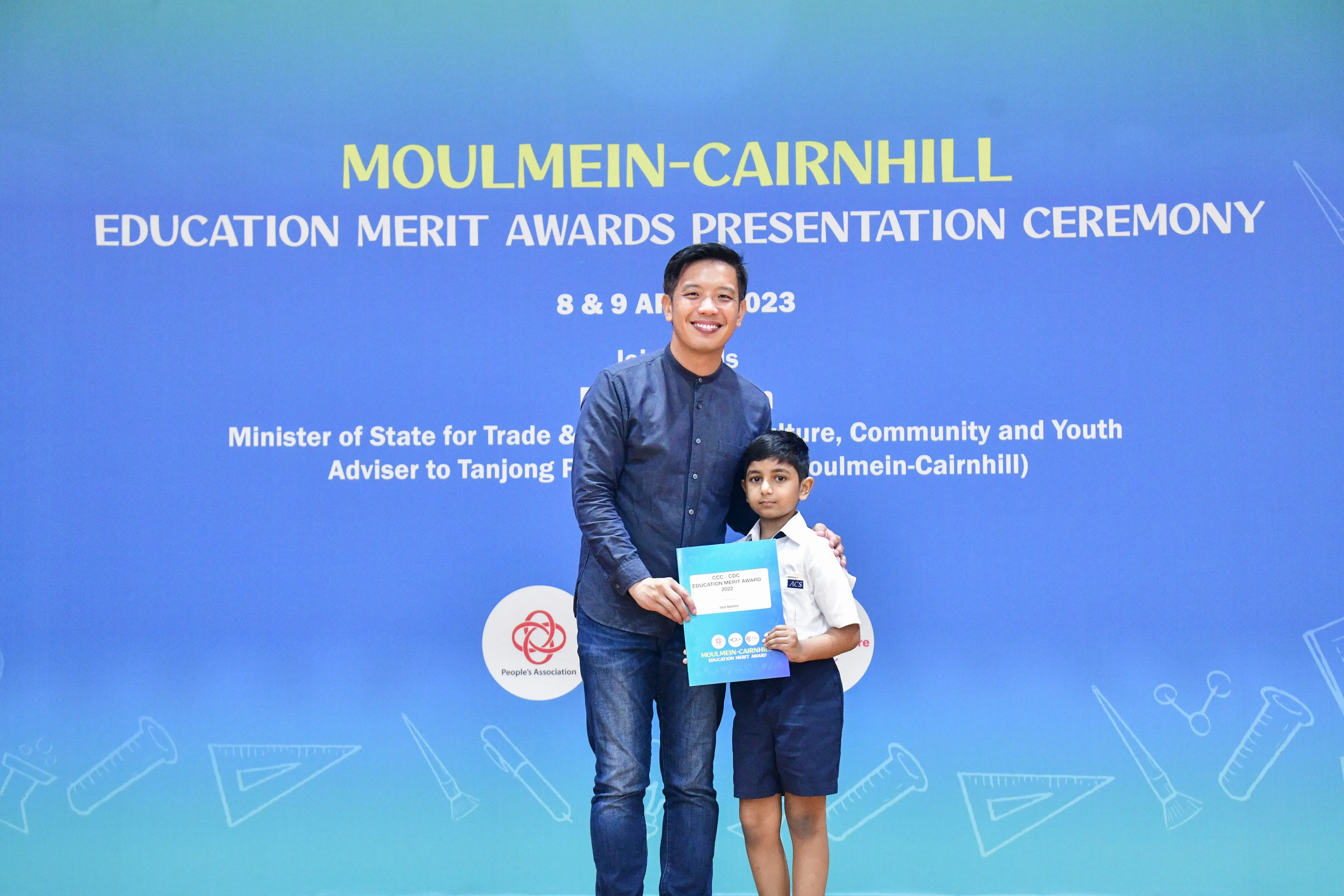 An adult and a young boy hold an award certificate in front of a Moulmein-Cairnhill Education Merit Awards banner.
