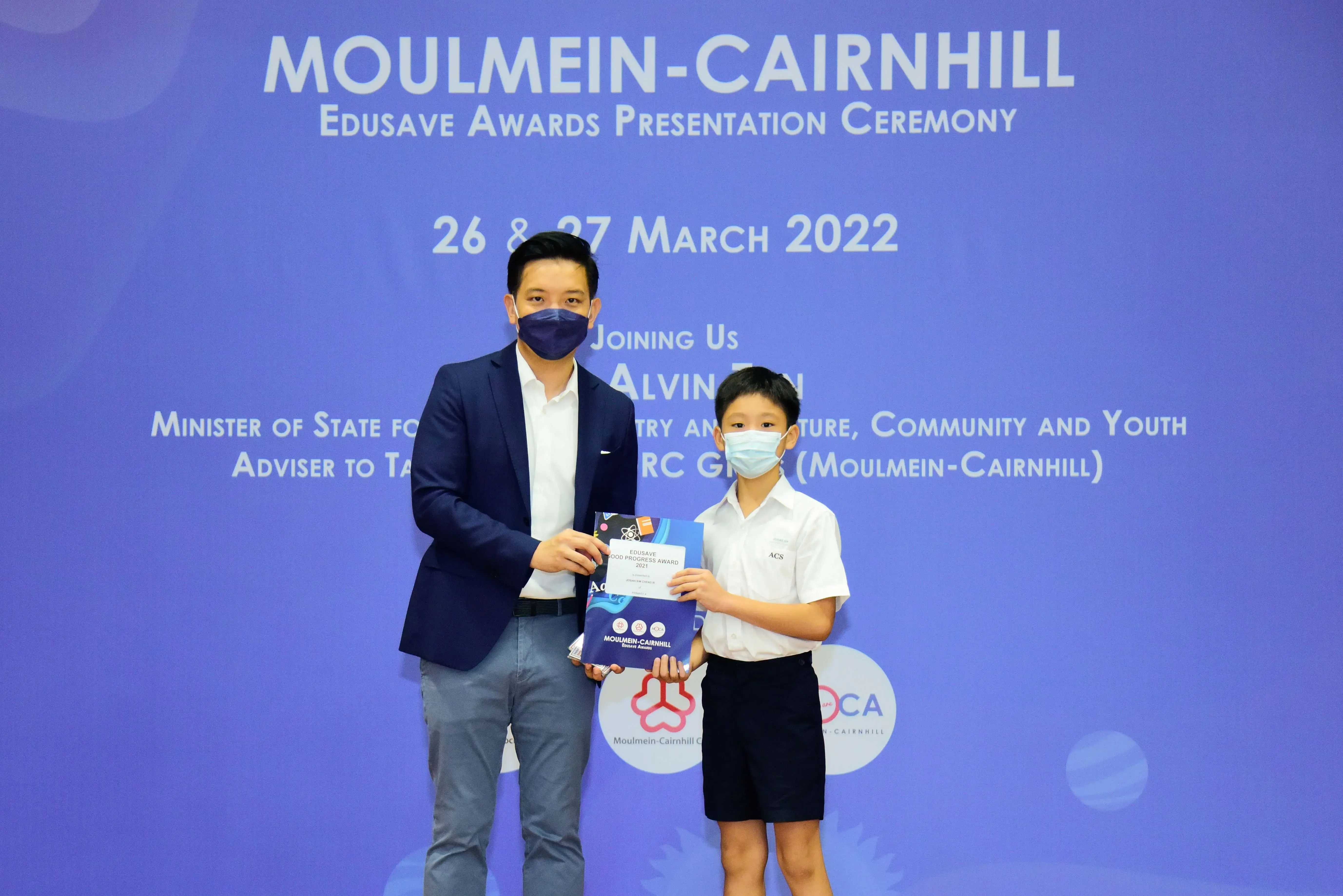 An adult and a child in masks hold an award certificate at the Moulmein-Cairnhill Edusave Awards Ceremony, March 2022.
