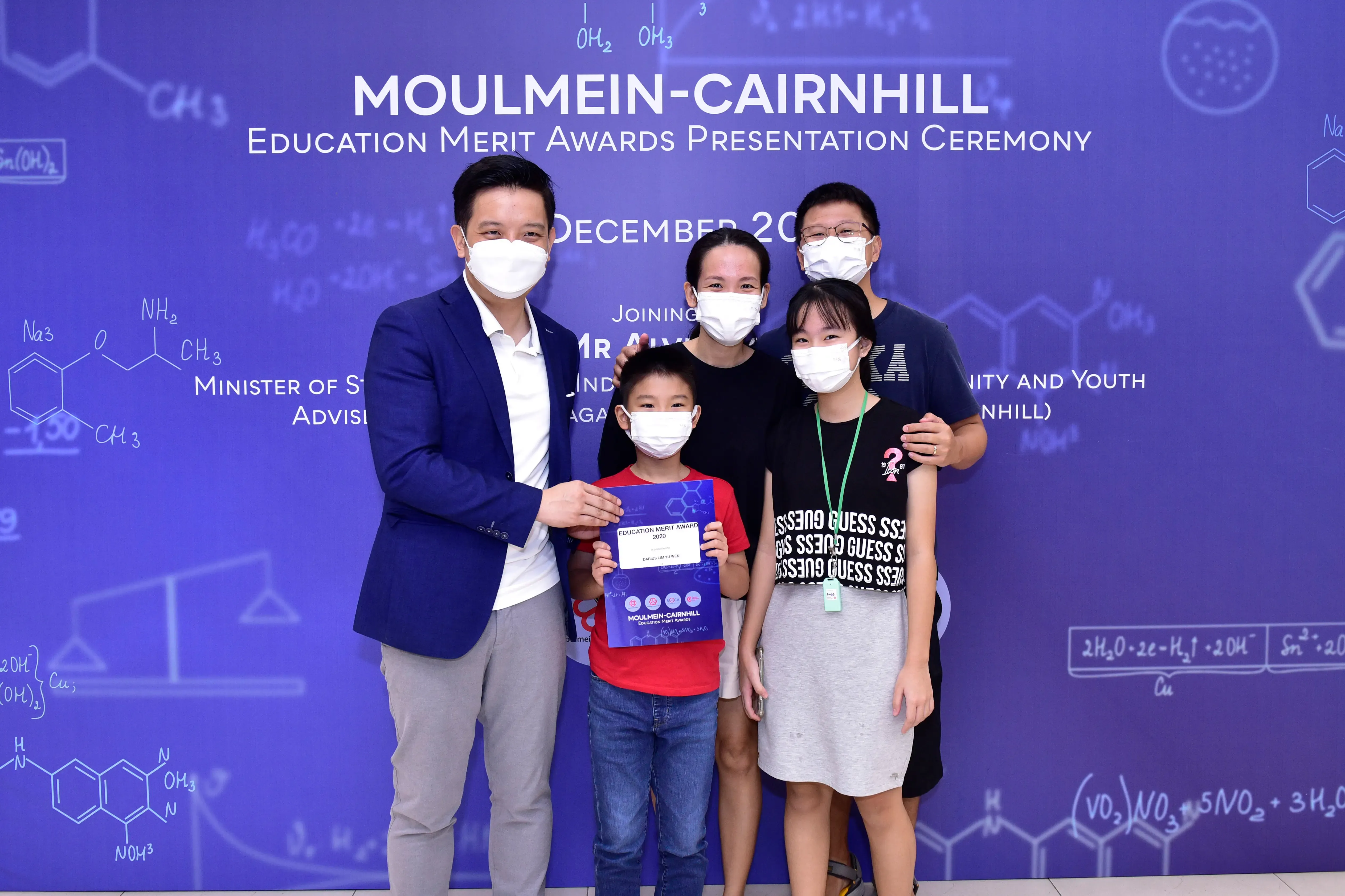 Five masked people pose at the Moulmein-Cairnhill Education Merit Awards Ceremony, with a boy holding a certificate.