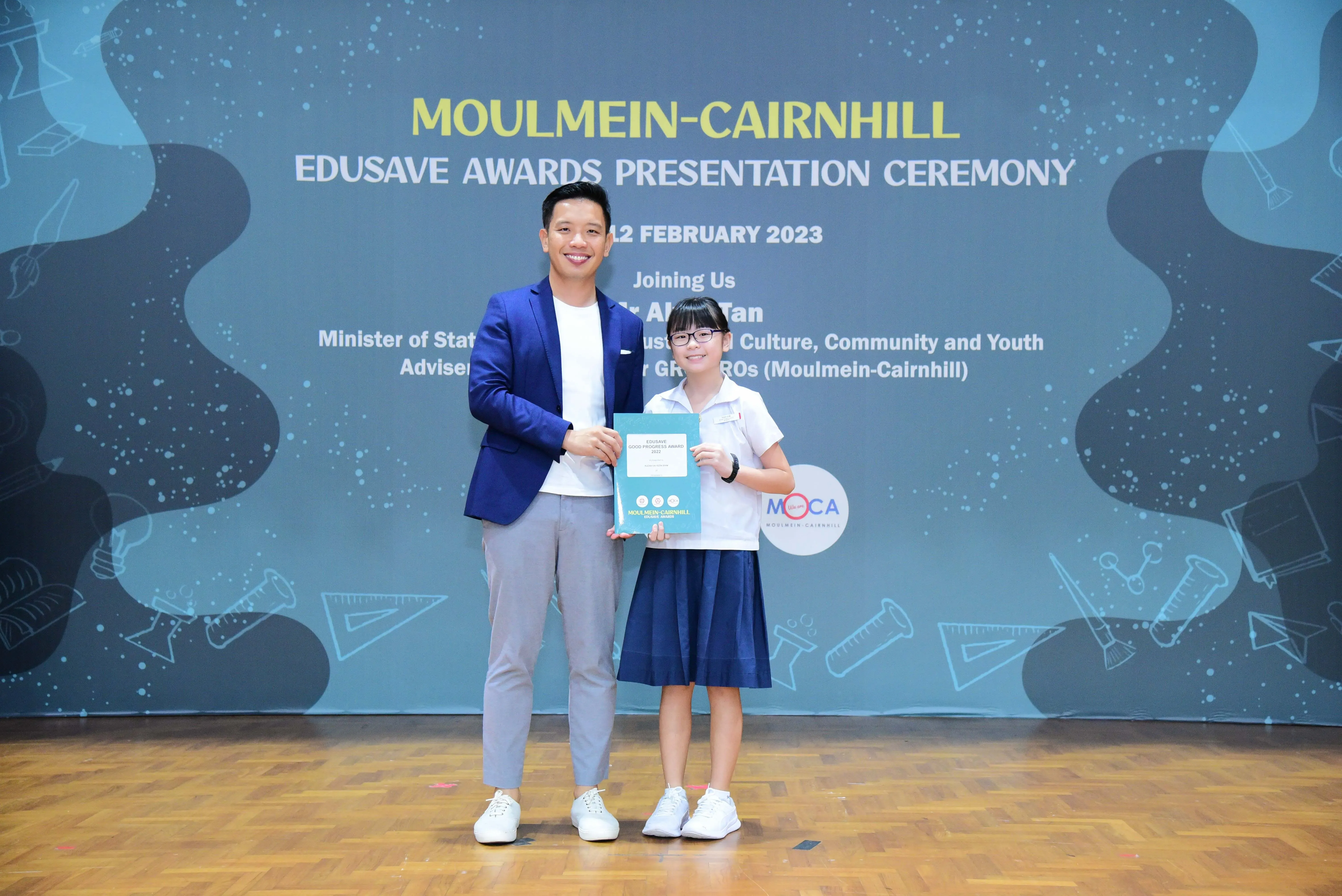 A man in a blue blazer and a young girl in school uniform hold a certificate at the Moulmein-Cairnhill Edusave Awards 2023.