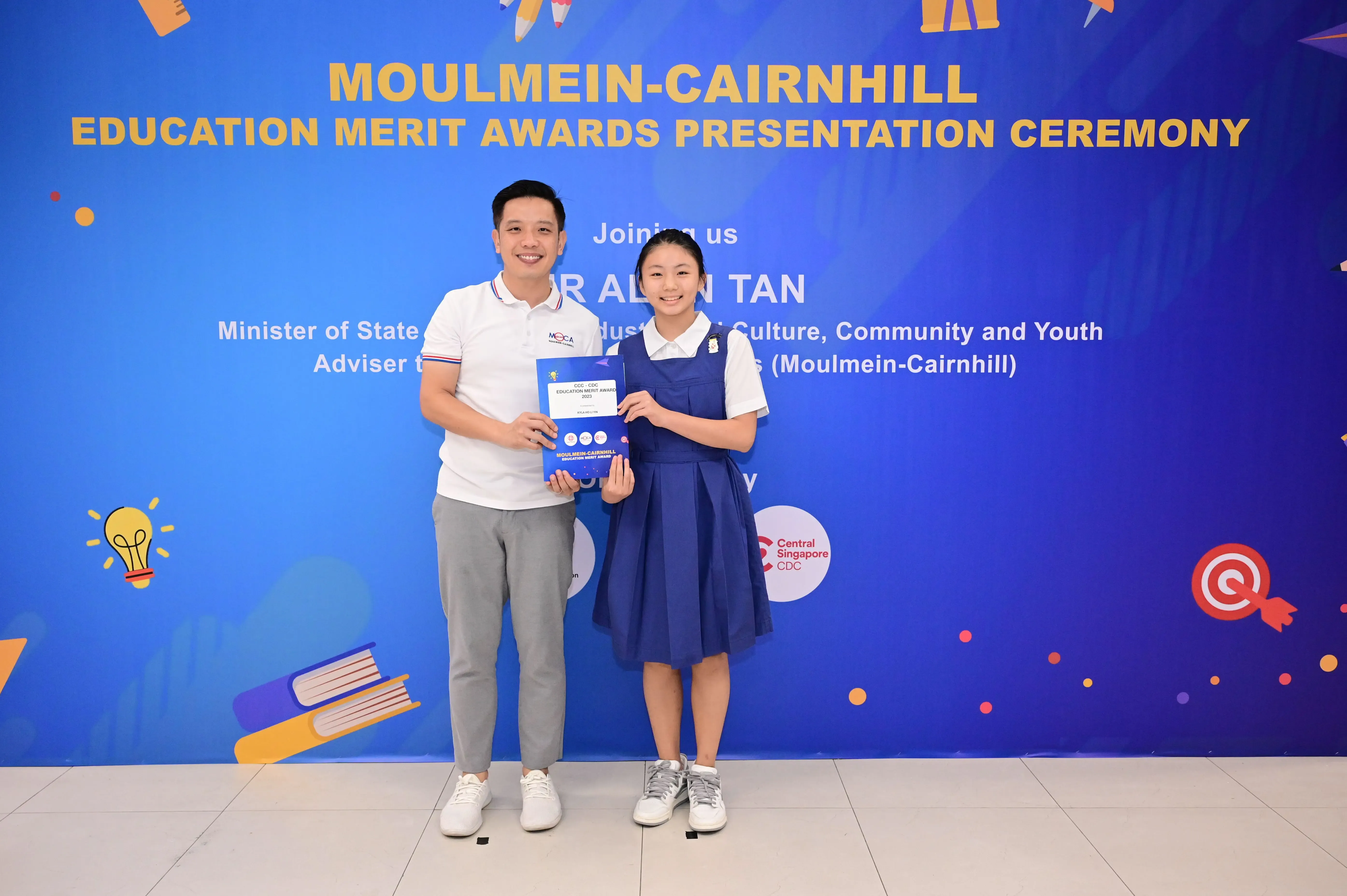 A man and a young girl hold an award certificate in front of a Moulmein-Cairnhill Education Merit Awards banner.