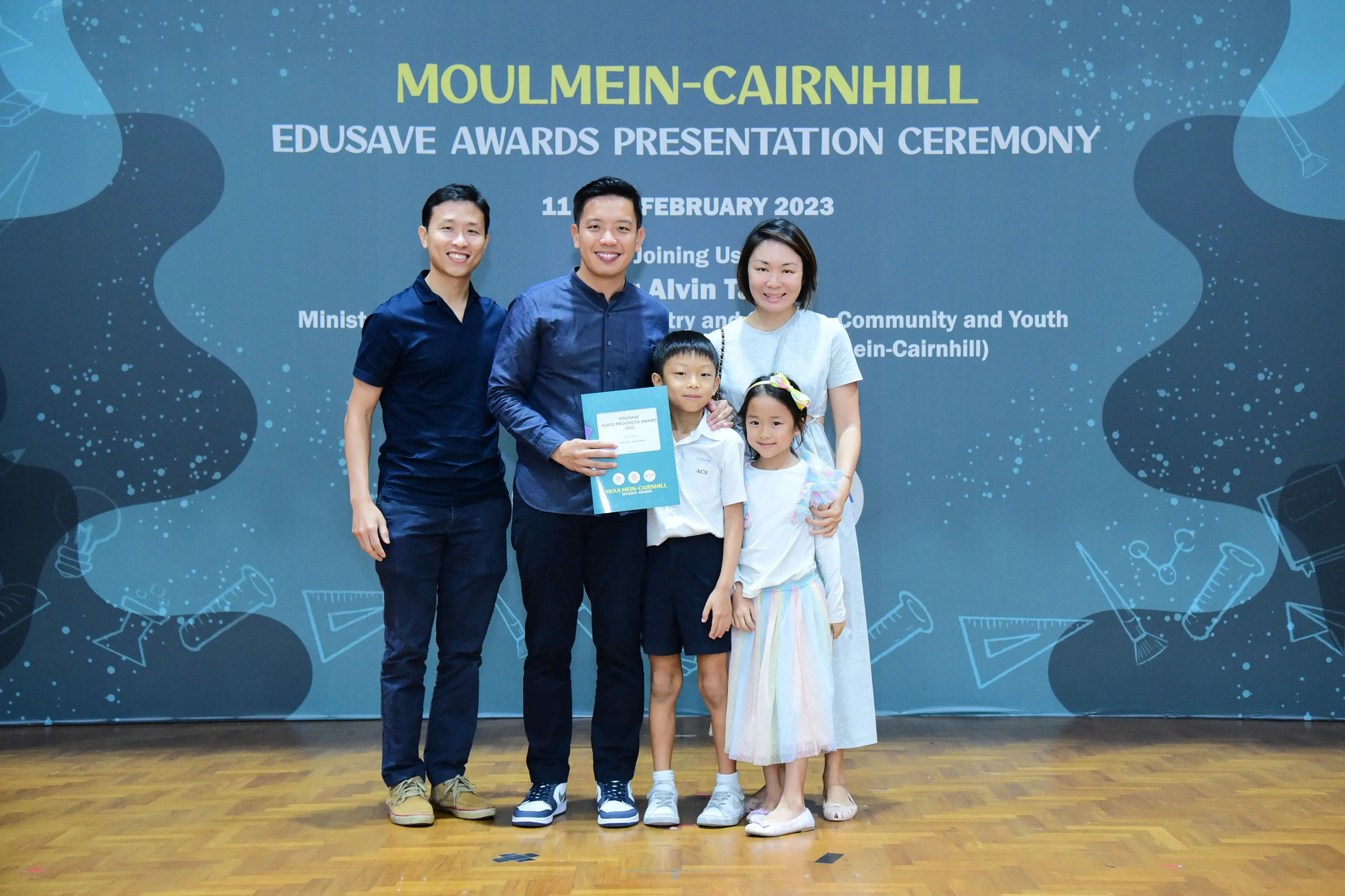 Four adults and two children pose with a certificate at the Moulmein-Cairnhill Edusave Awards Presentation Ceremony 2023.