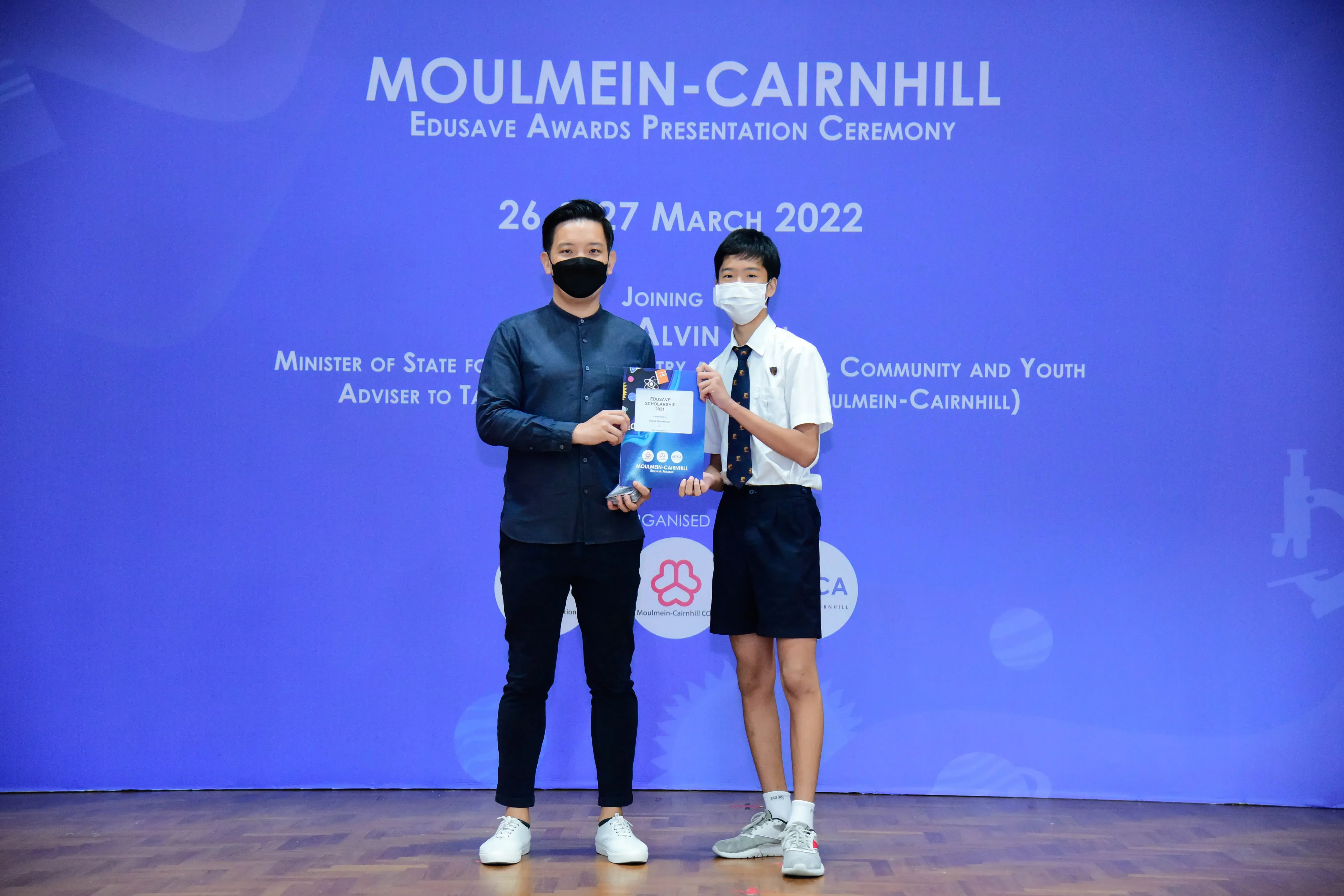 Two masked individuals hold an award certificate on stage at the Moulmein-Cairnhill Edusave Awards Ceremony, March 2022.