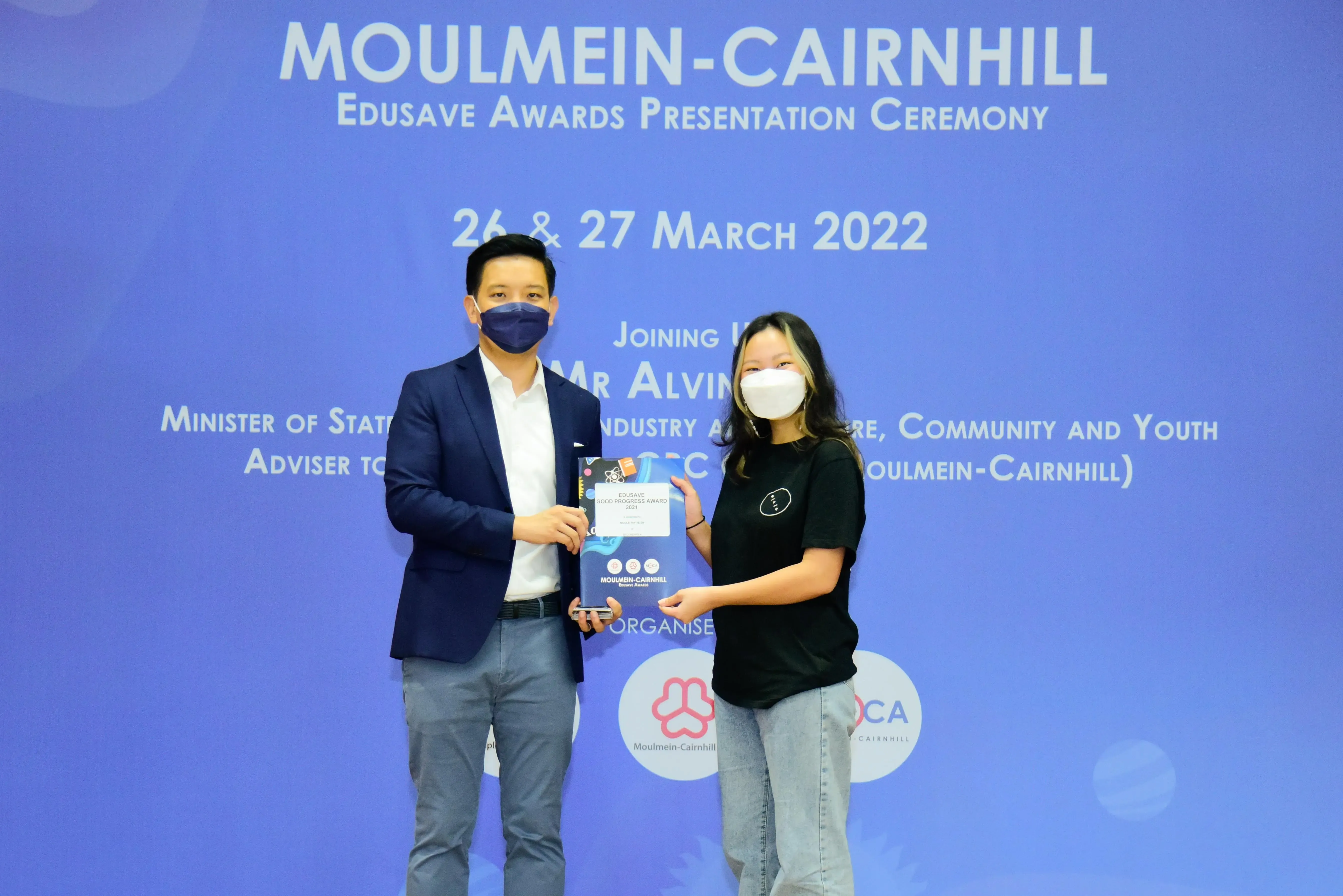 Two masked people hold an award at the Moulmein-Cairnhill Edusave Awards Presentation Ceremony, March 2022.