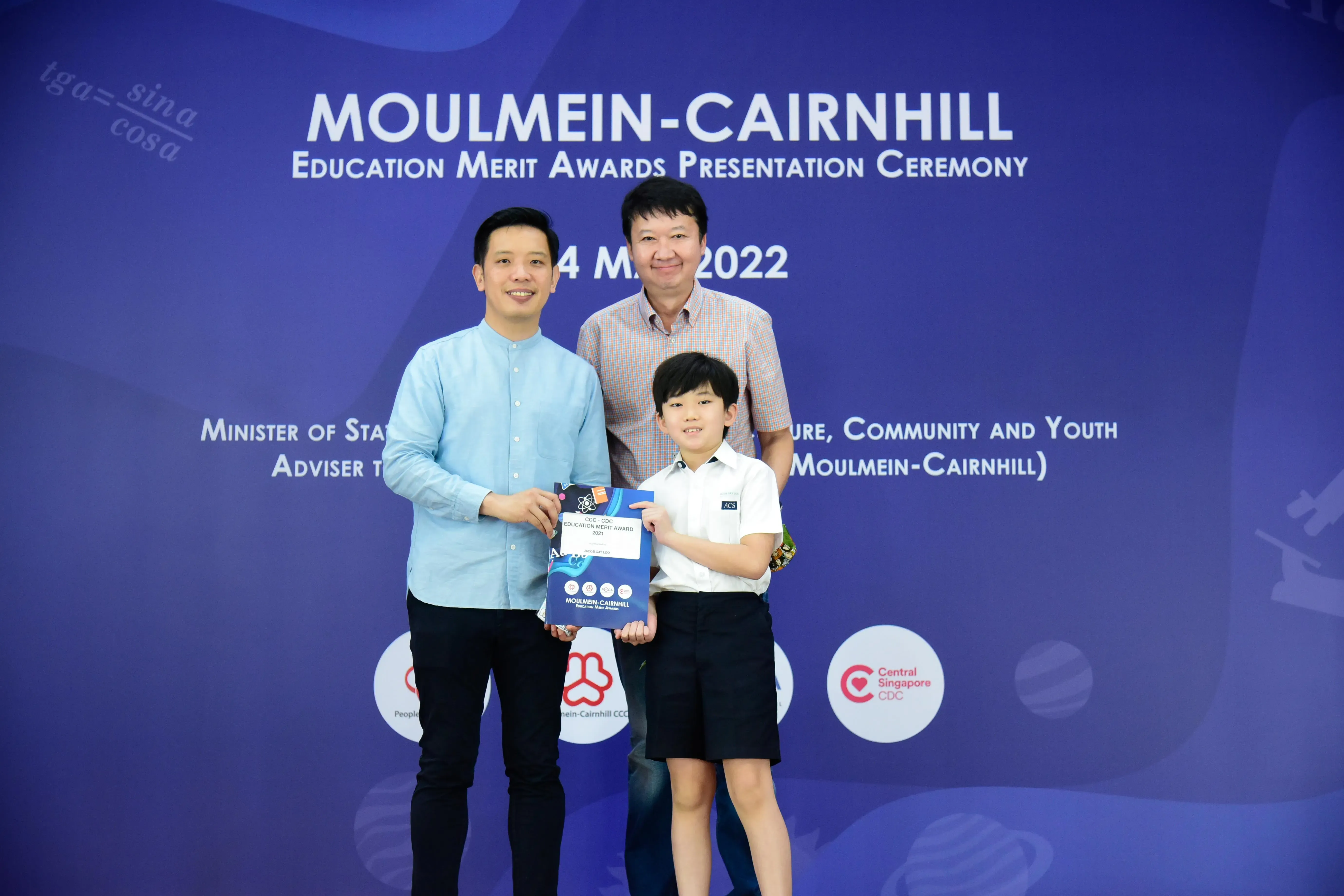 Two men and a young boy hold an award certificate at the Moulmein-Cairnhill Education Merit Awards Ceremony 2022.