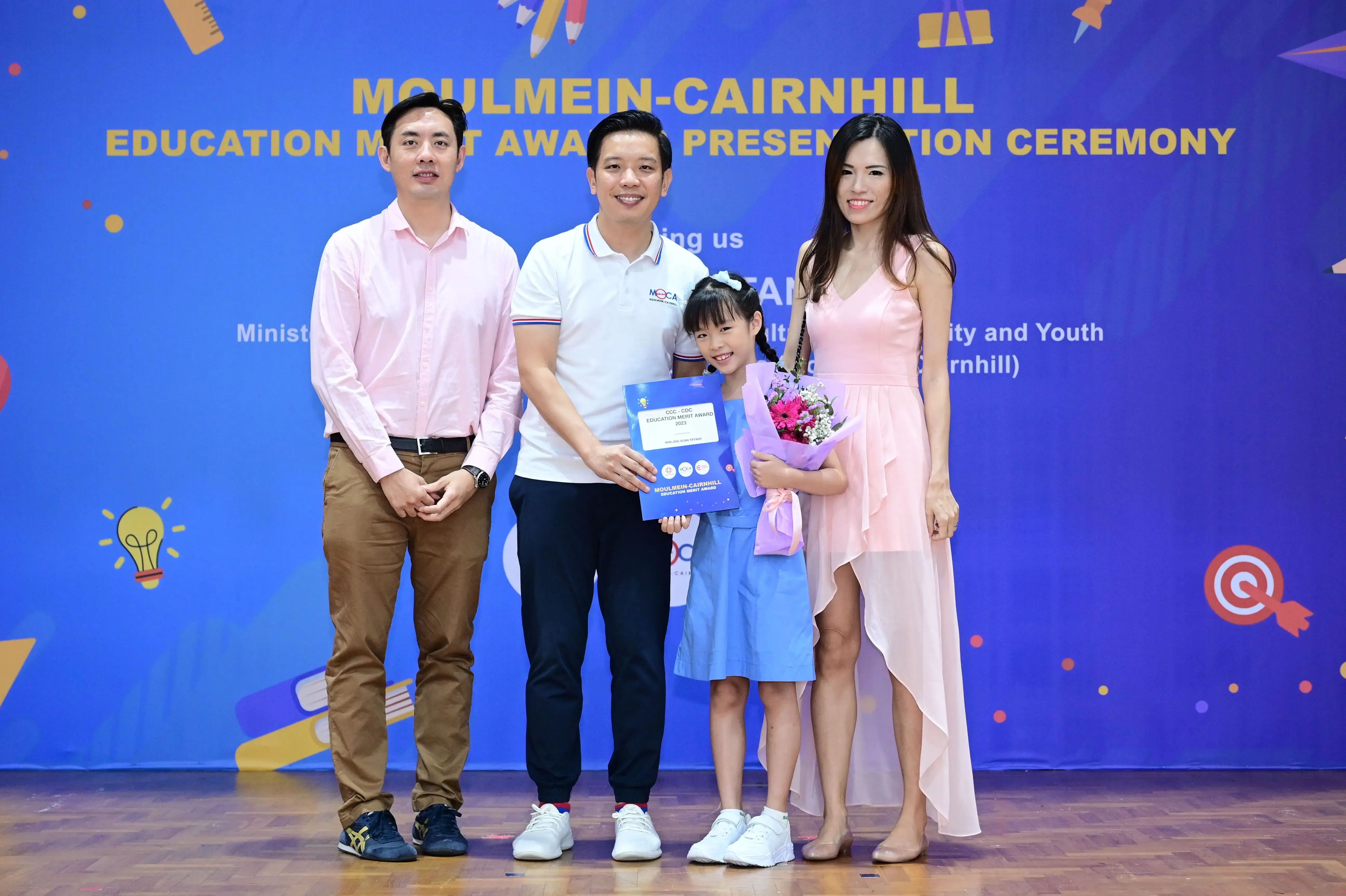 Three adults and a young girl pose at the Moulmein-Cairnhill Education Merit Award Presentation Ceremony, girl holds flowers and a certificate.