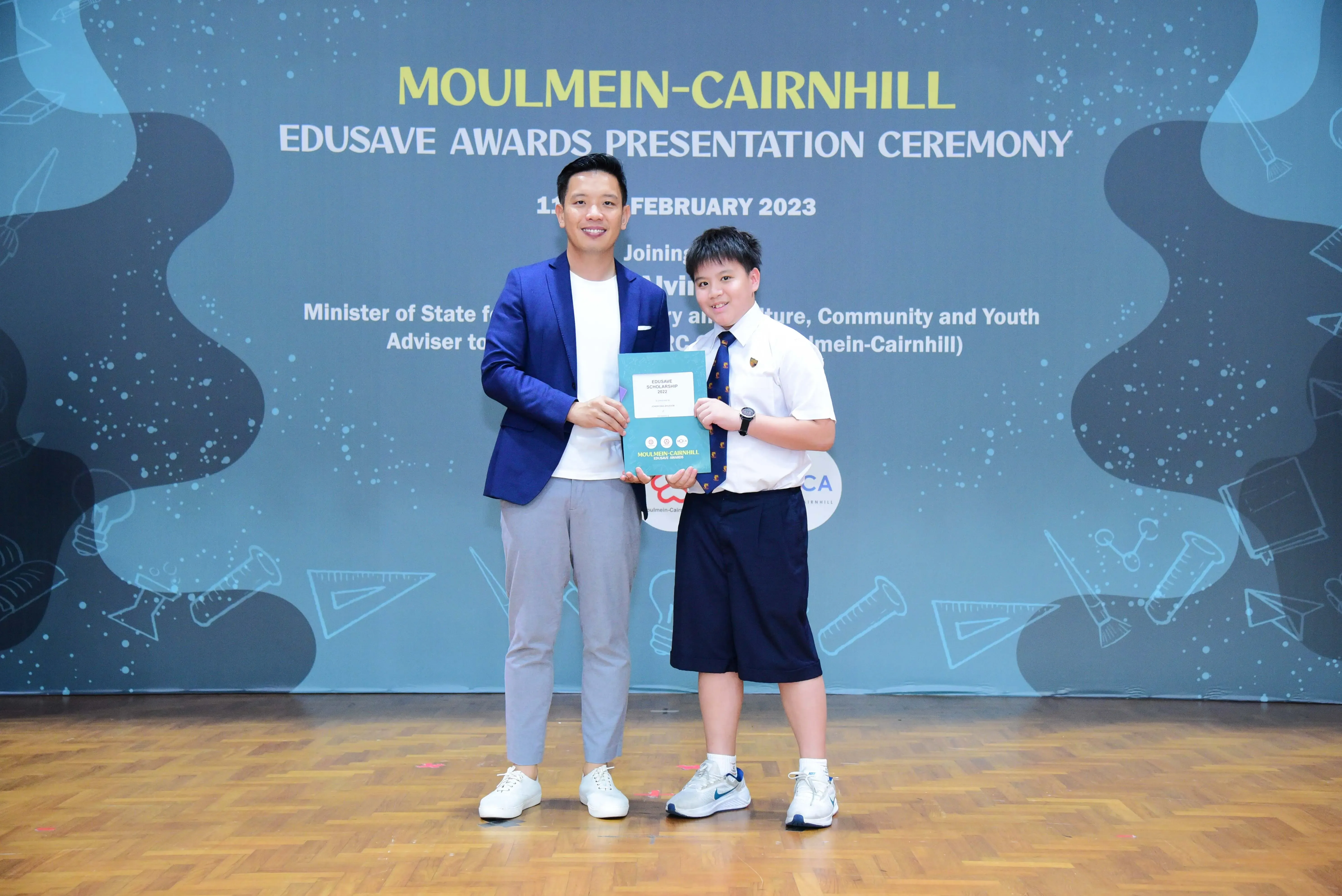 An adult in a blue blazer and a uniformed student hold an award certificate at the Moulmein-Cairnhill Edusave Awards Ceremony.