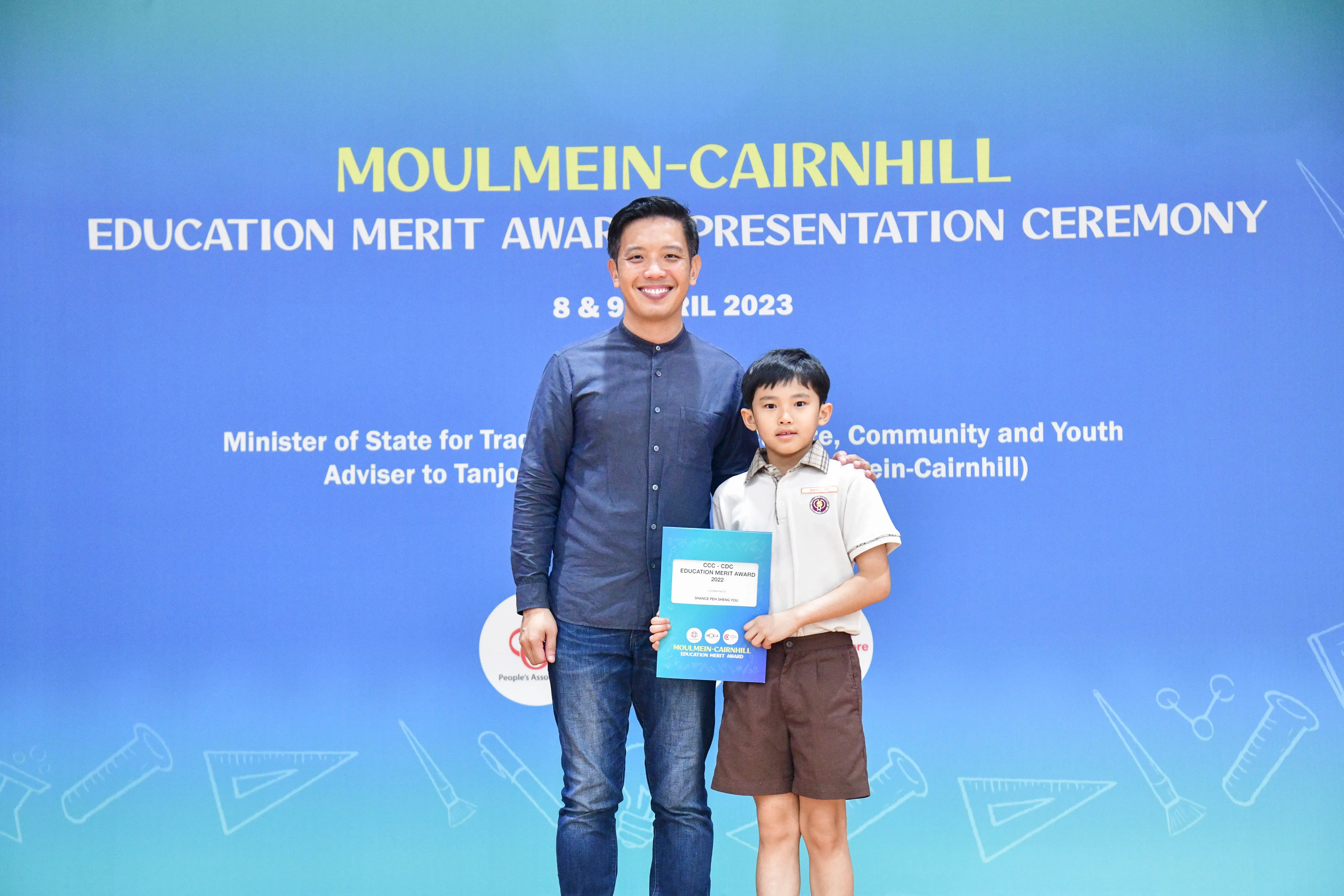 An adult man and a young boy holding an award certificate stand before a Moulmein-Cairnhill Education Merit Award 2023 banner.