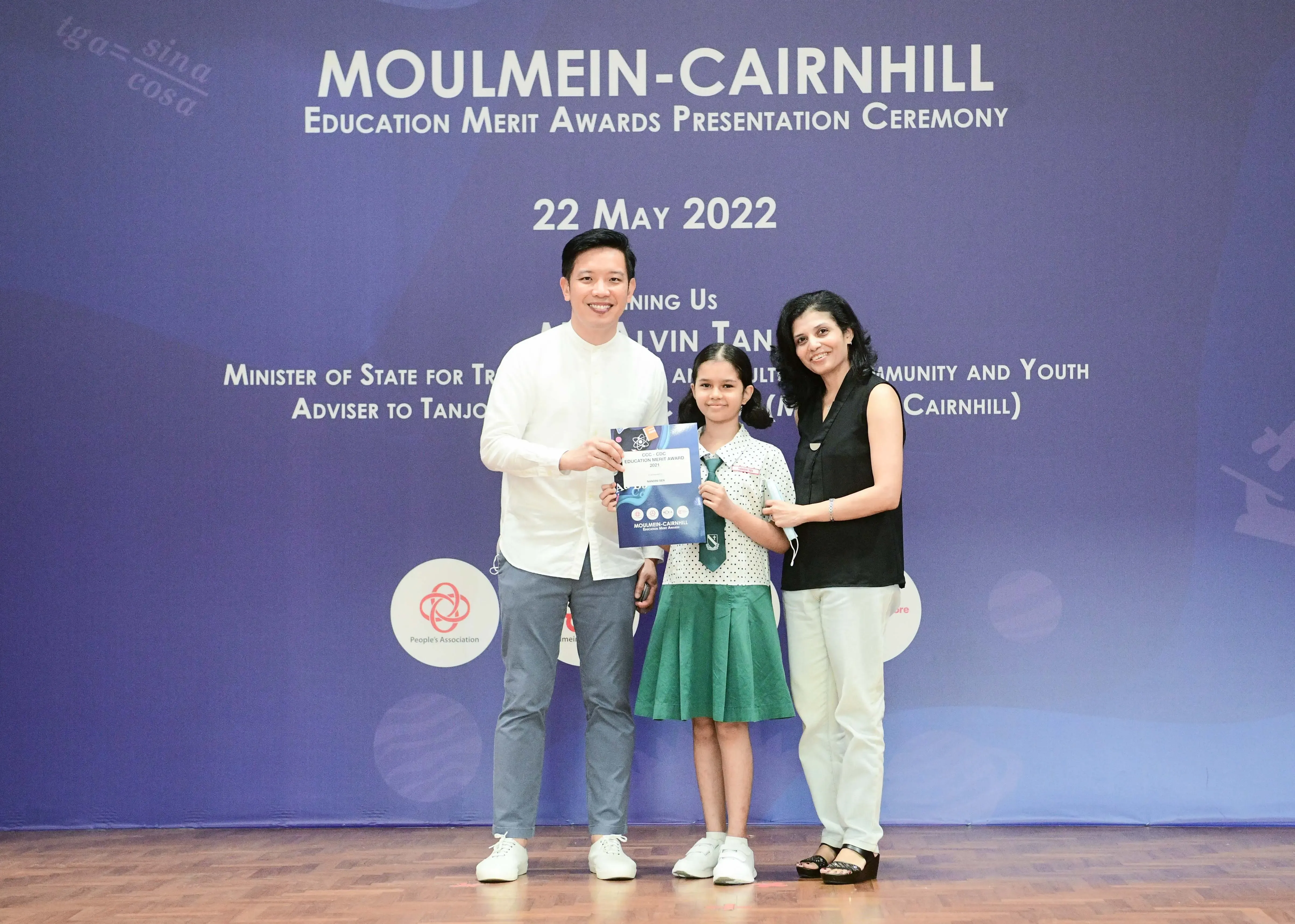 A girl in school uniform holds an award certificate flanked by two adults at the Moulmein-Cairnhill Education Merit Awards 2022.