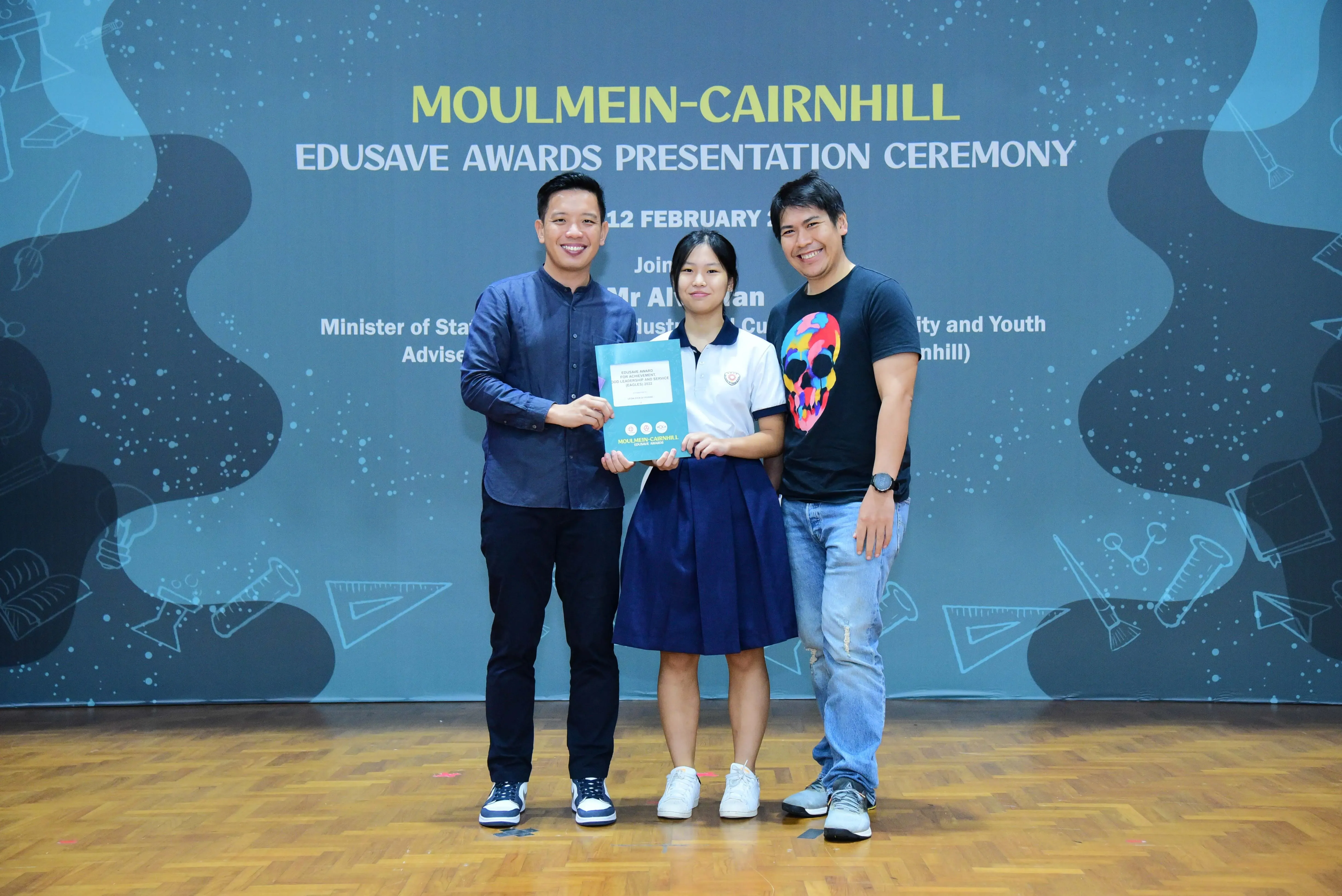 Three people pose holding an award folder at the Moulmein-Cairnhill Edusave Awards Presentation Ceremony backdrop.