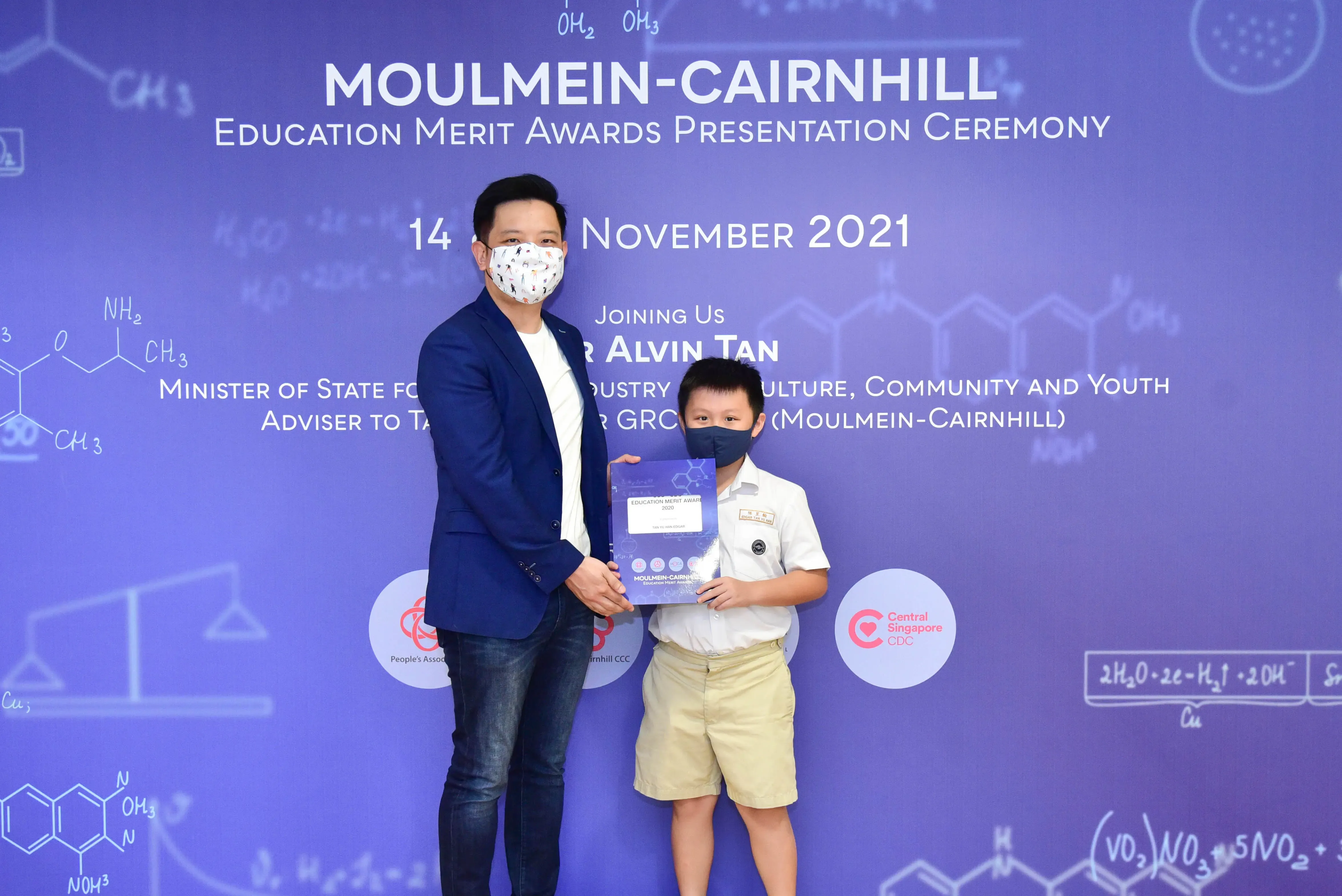 An adult and a young boy wearing masks hold a certificate at the Moulmein-Cairnhill Education Merit Awards 2021.
