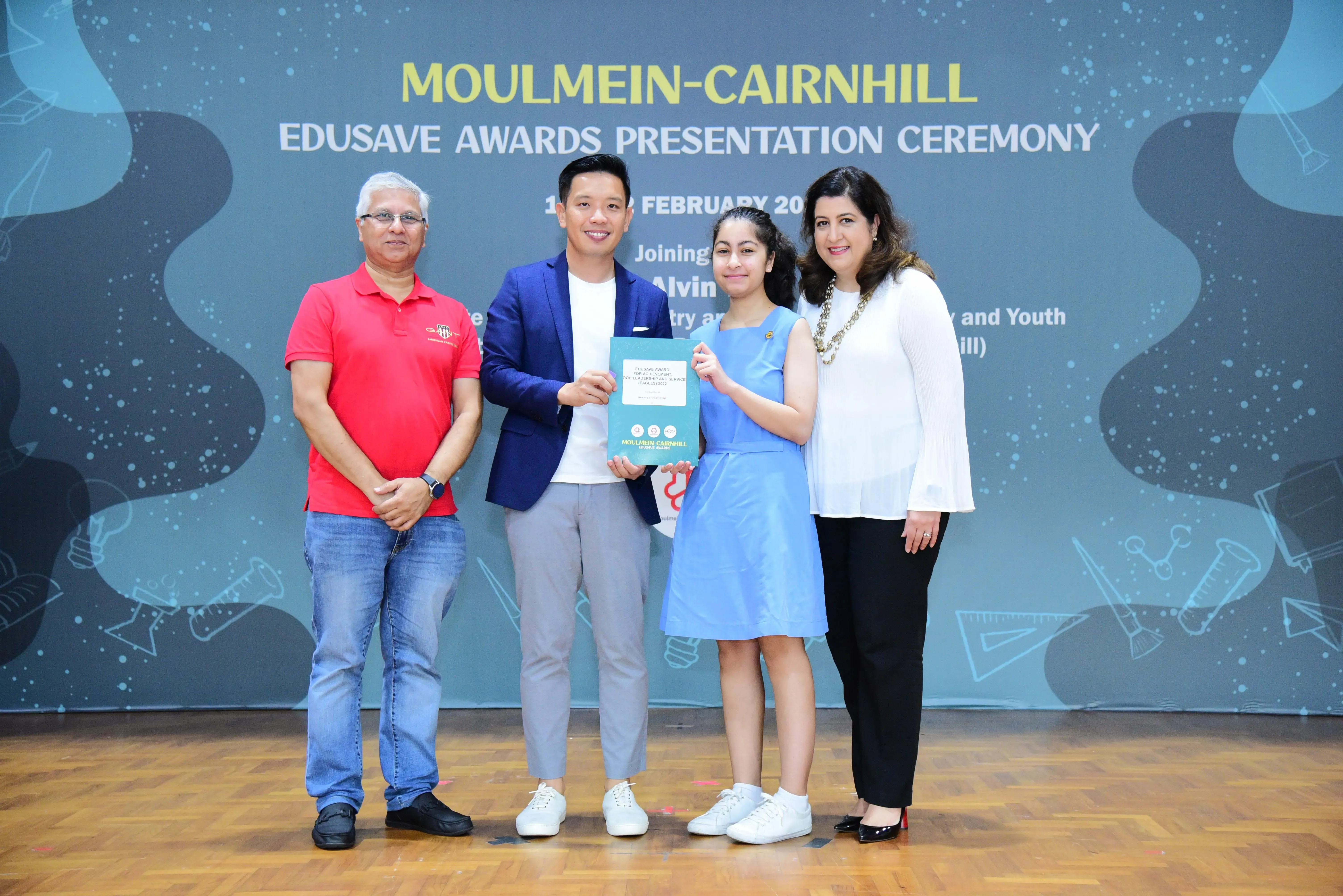 Four people pose on a stage at the Moulmein-Cairnhill Edusave Awards Presentation Ceremony, holding a certificate.