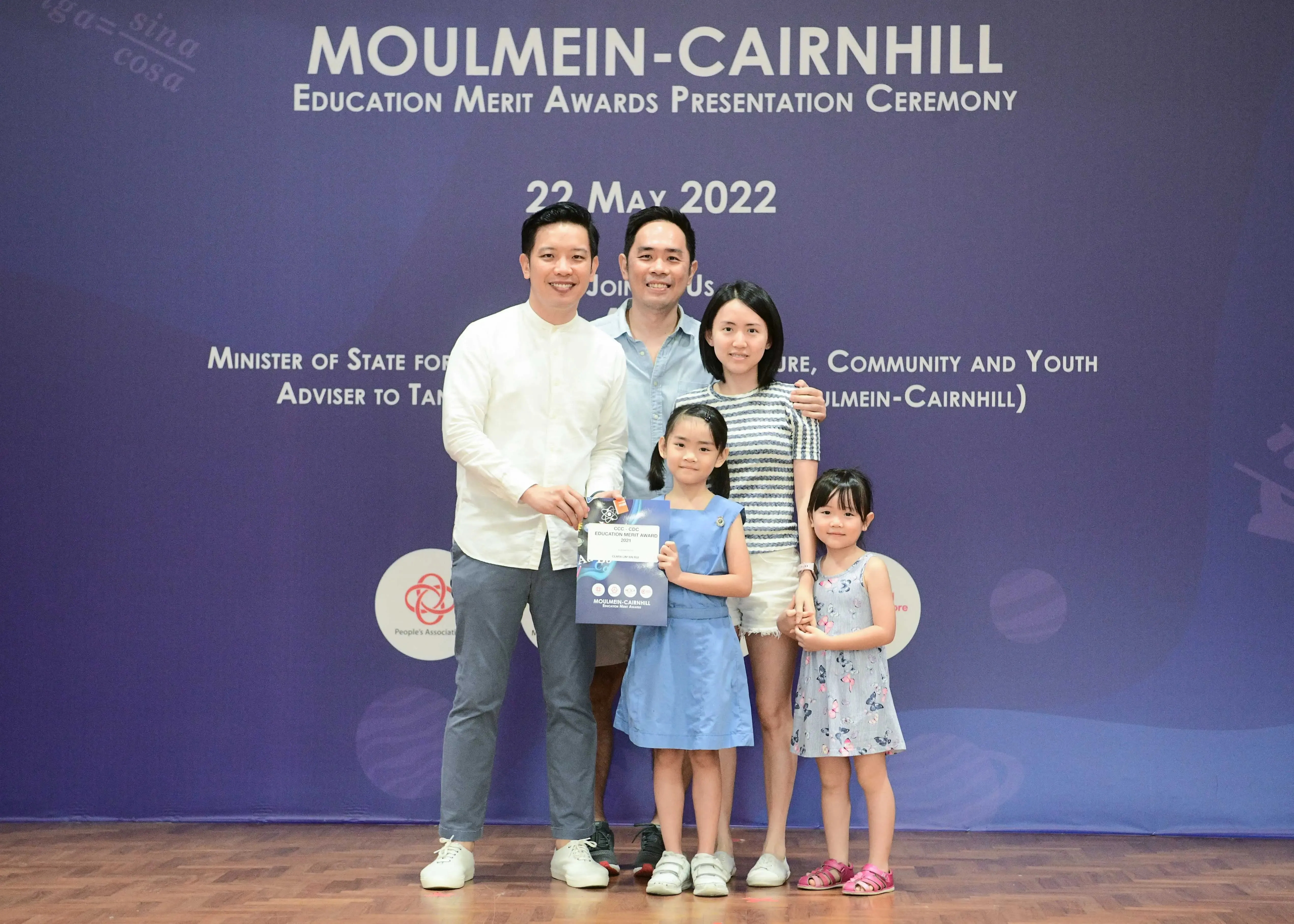 Five people pose before a Moulmein-Cairnhill Education Merit Awards banner dated 22 May 2022, with a girl holding a certificate.