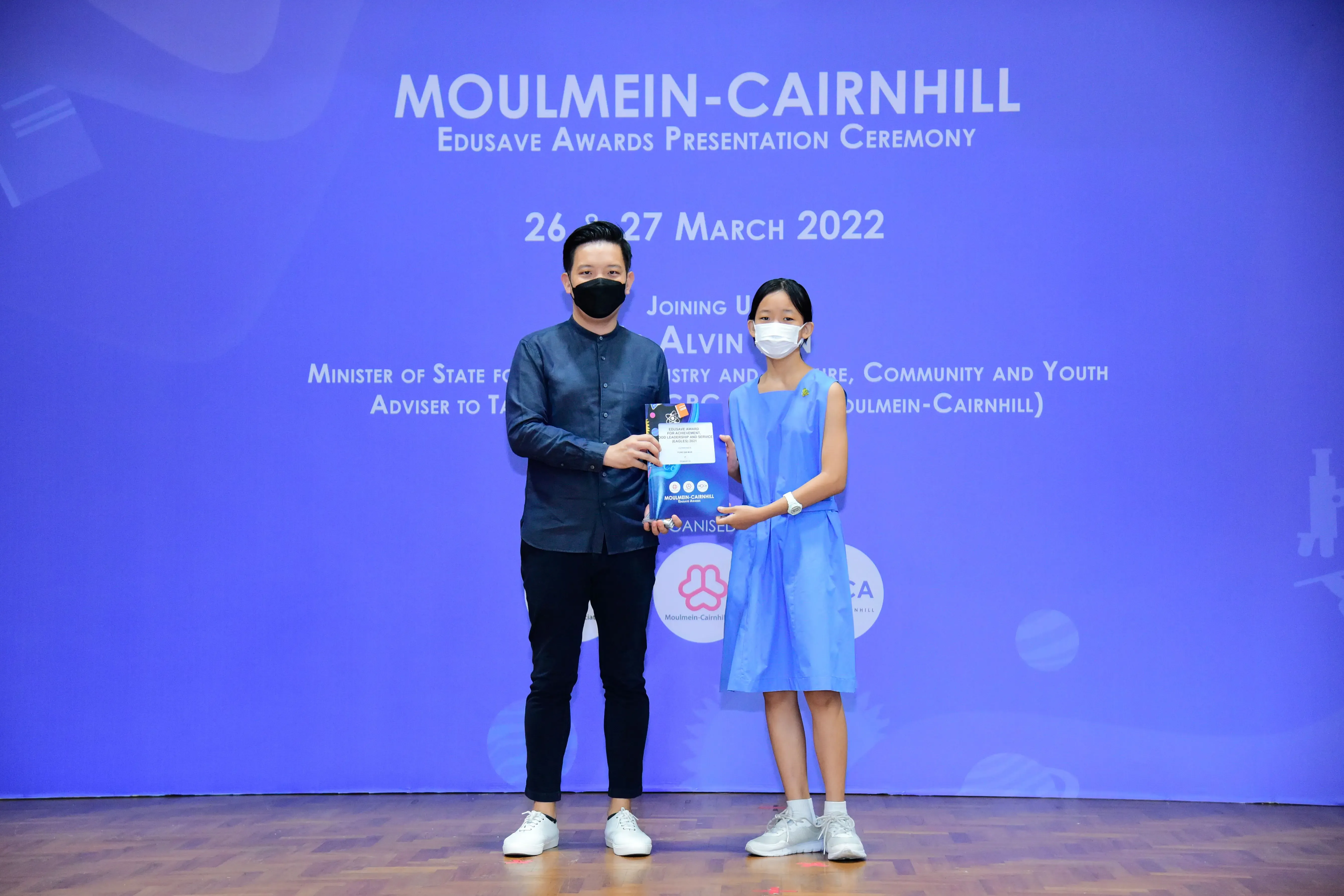An adult and a young girl in masks hold an award certificate at the Moulmein-Cairnhill Edusave Awards Ceremony 2022.