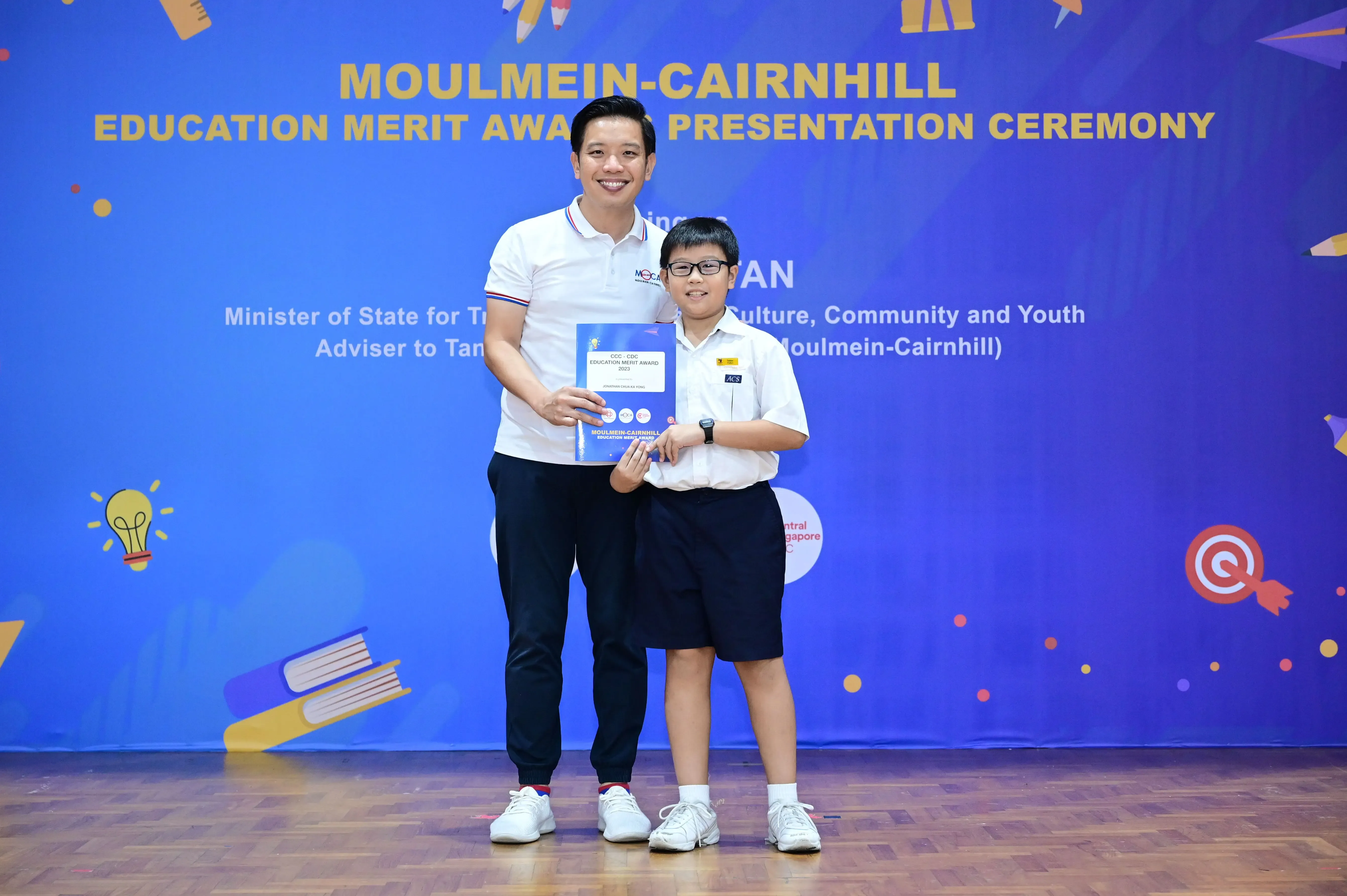 An adult and a young student hold an award certificate at the Moulmein-Cairnhill Education Merit Award Presentation Ceremony.