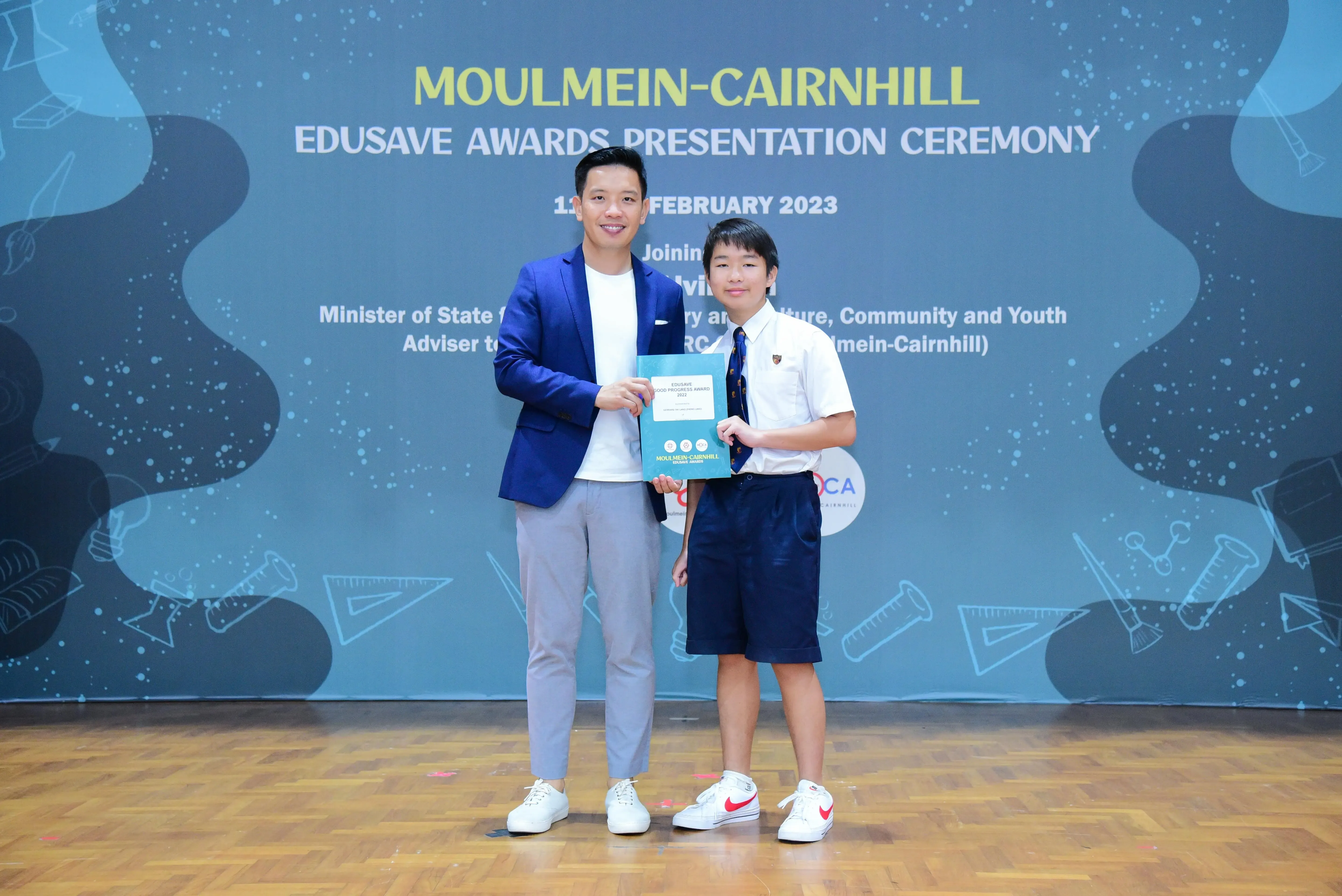 Adult in blue blazer and young student in uniform hold a certificate at Moulmein-Cairnhill Edusave Awards Ceremony 2023.