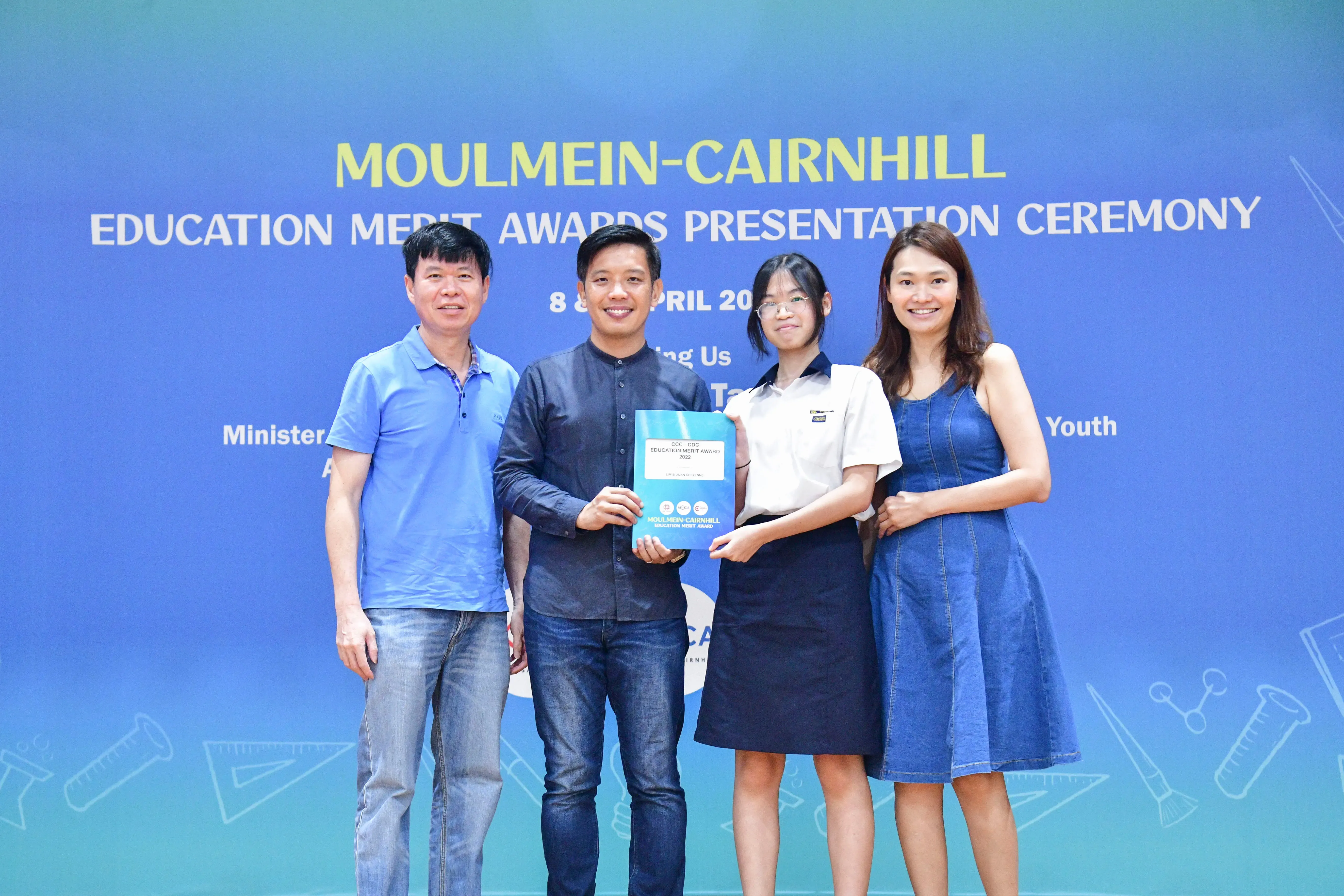Four people pose holding an award certificate in front of a Moulmein-Cairnhill Education Merit Awards banner.