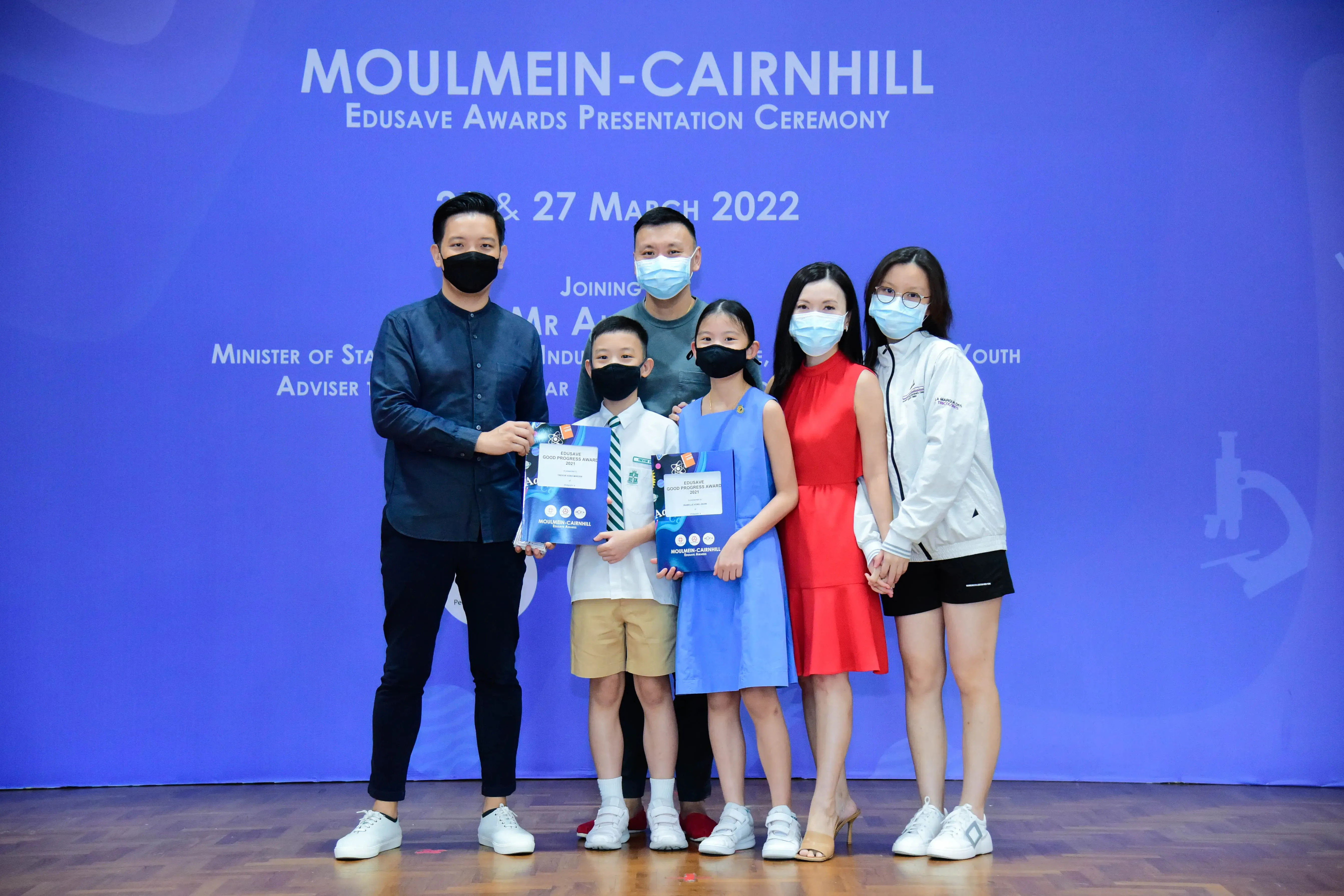 Six masked people pose at the Moulmein-Cairnhill Edusave Awards Ceremony in March 2022, two children holding award folders.