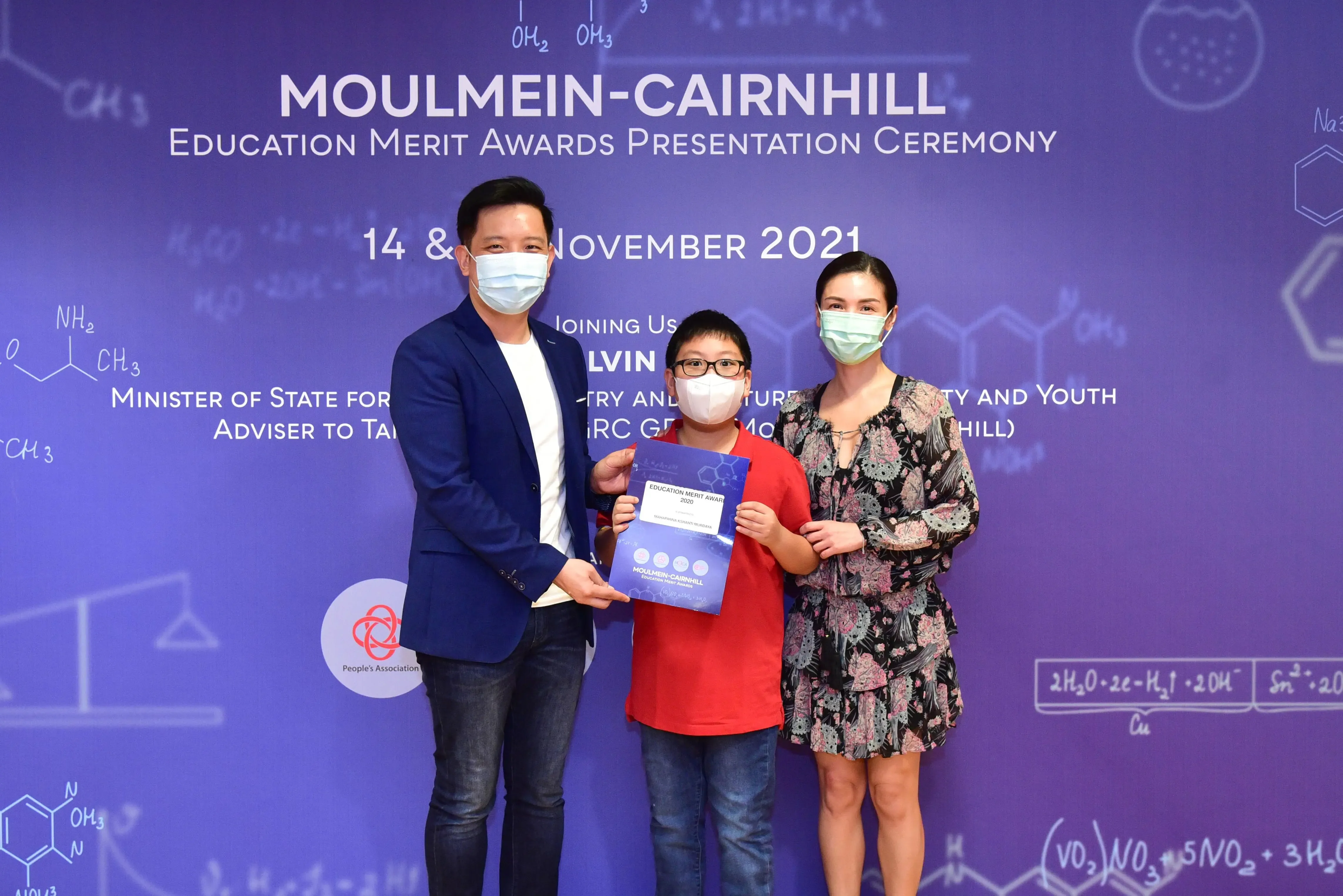 Three masked people pose at the Moulmein-Cairnhill Education Merit Awards Ceremony, November 2021, child holding a certificate.