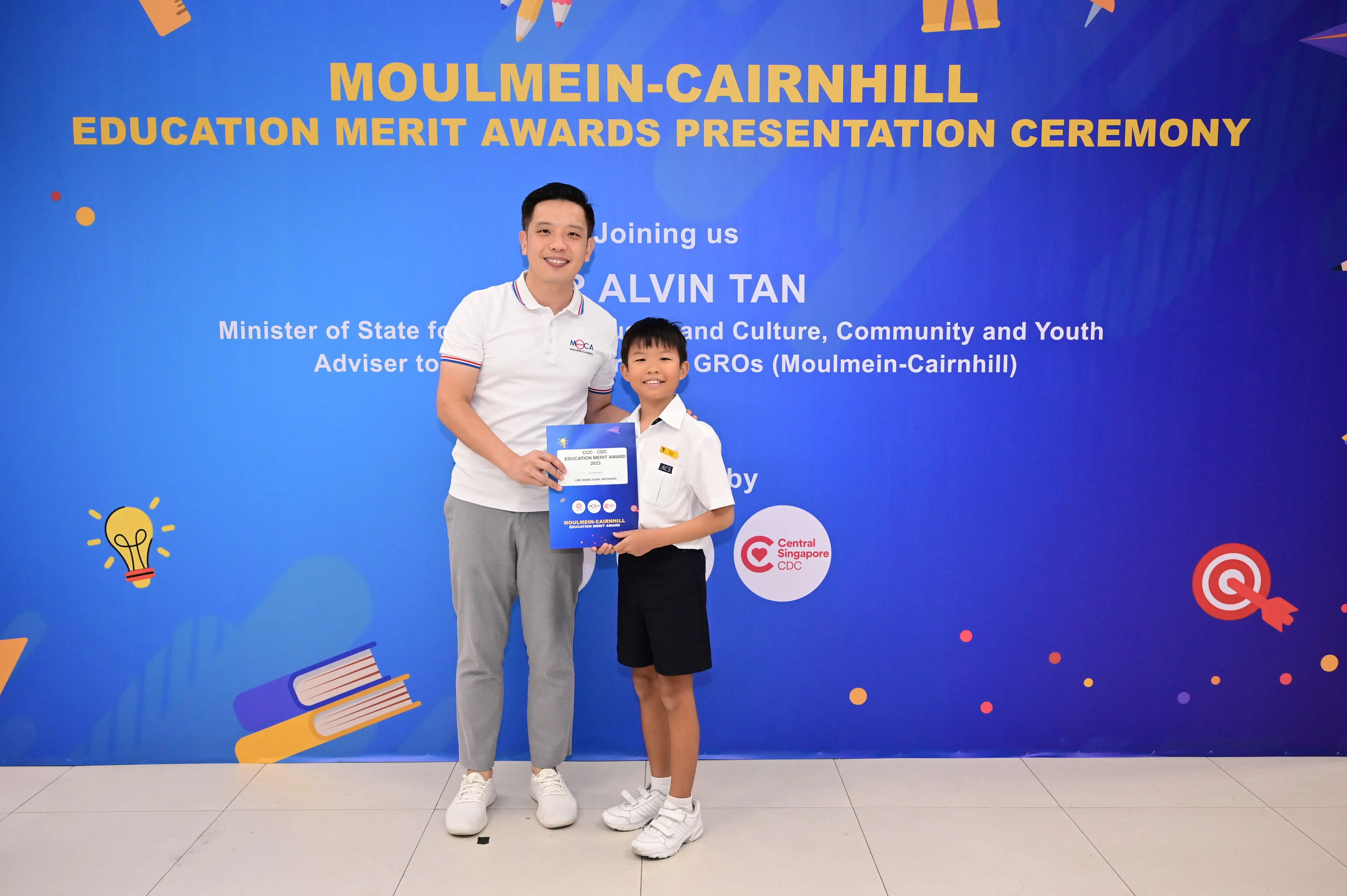 A man and a young boy hold award certificates before a Moulmein-Cairnhill Education Merit Awards banner.