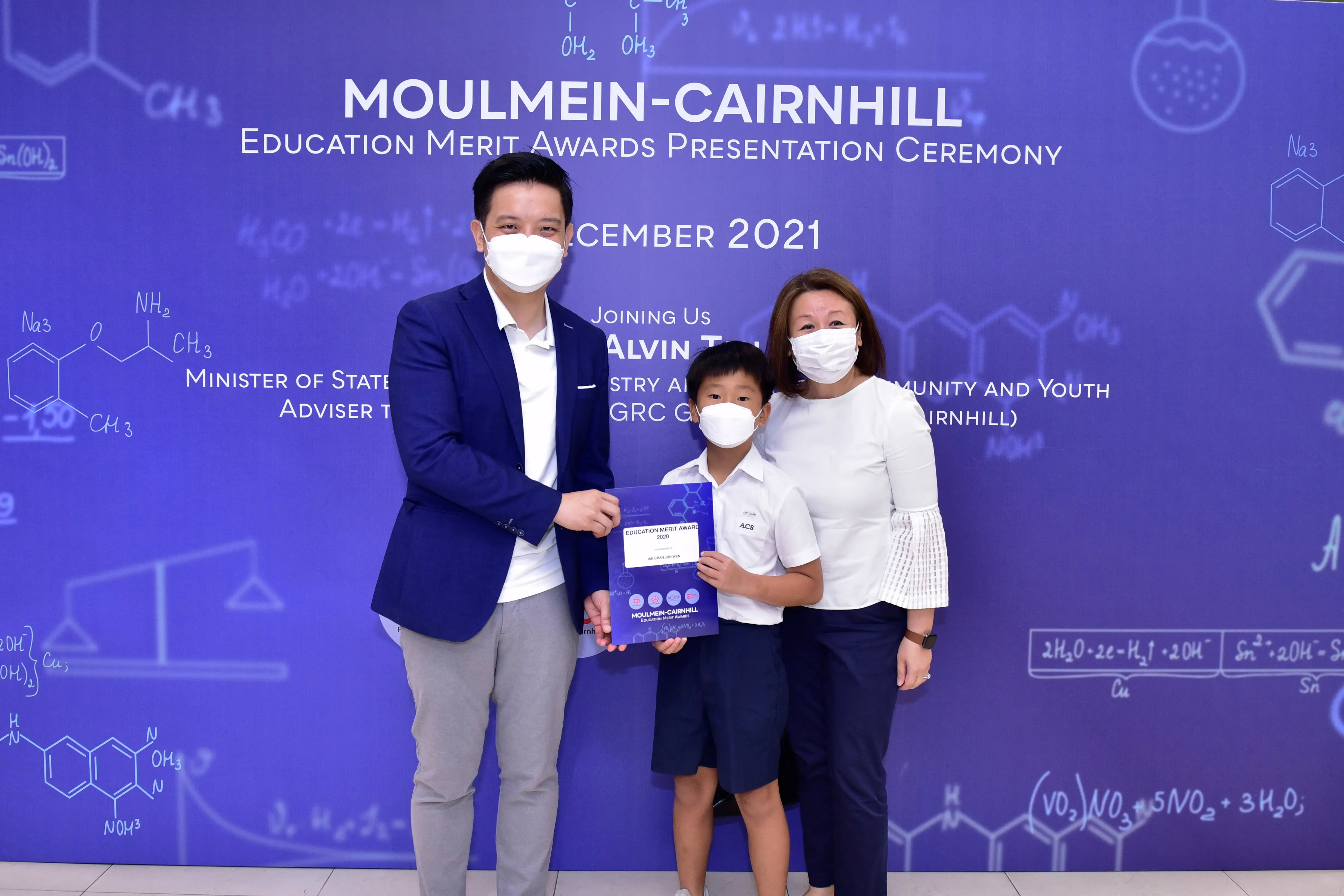 Two masked adults and a masked boy holding an award certificate at the Moulmein-Cairnhill Education Merit Awards 2021.