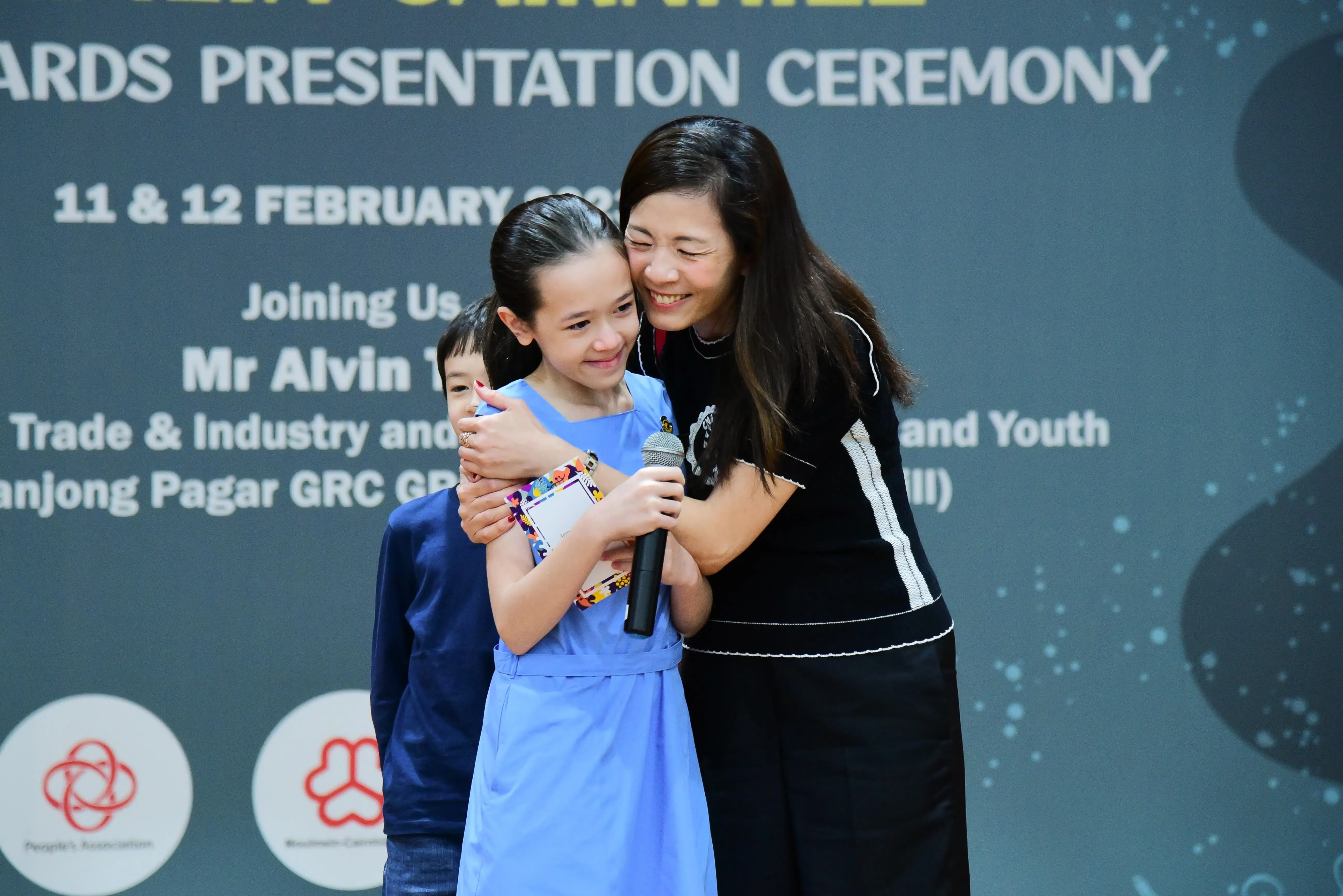 A woman hugs a smiling girl holding a microphone on stage at an awards presentation ceremony backdrop.