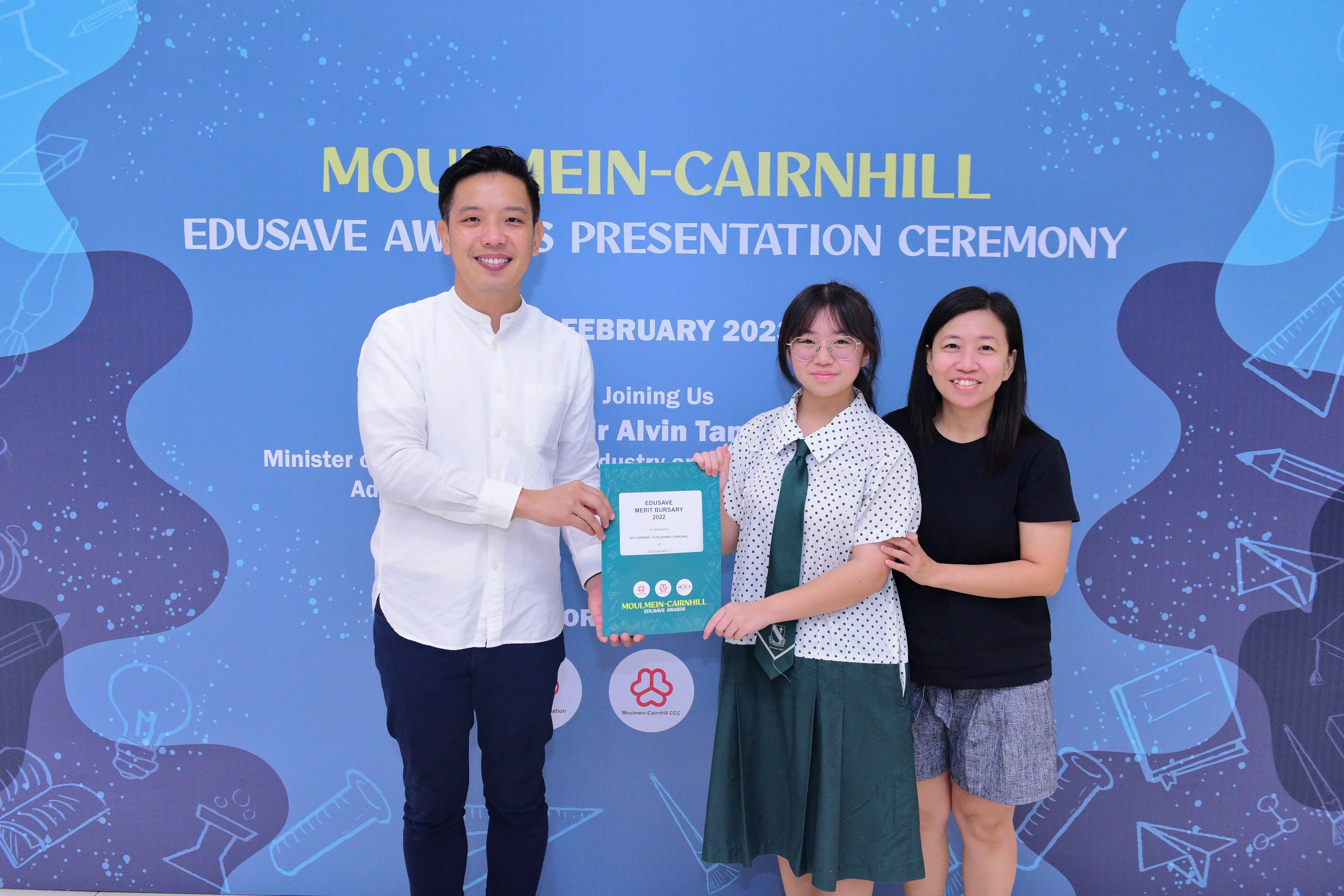 Three people smile holding an Edusave Merit Bursary certificate at the Moulmein-Cairnhill Edusave Awards Ceremony.