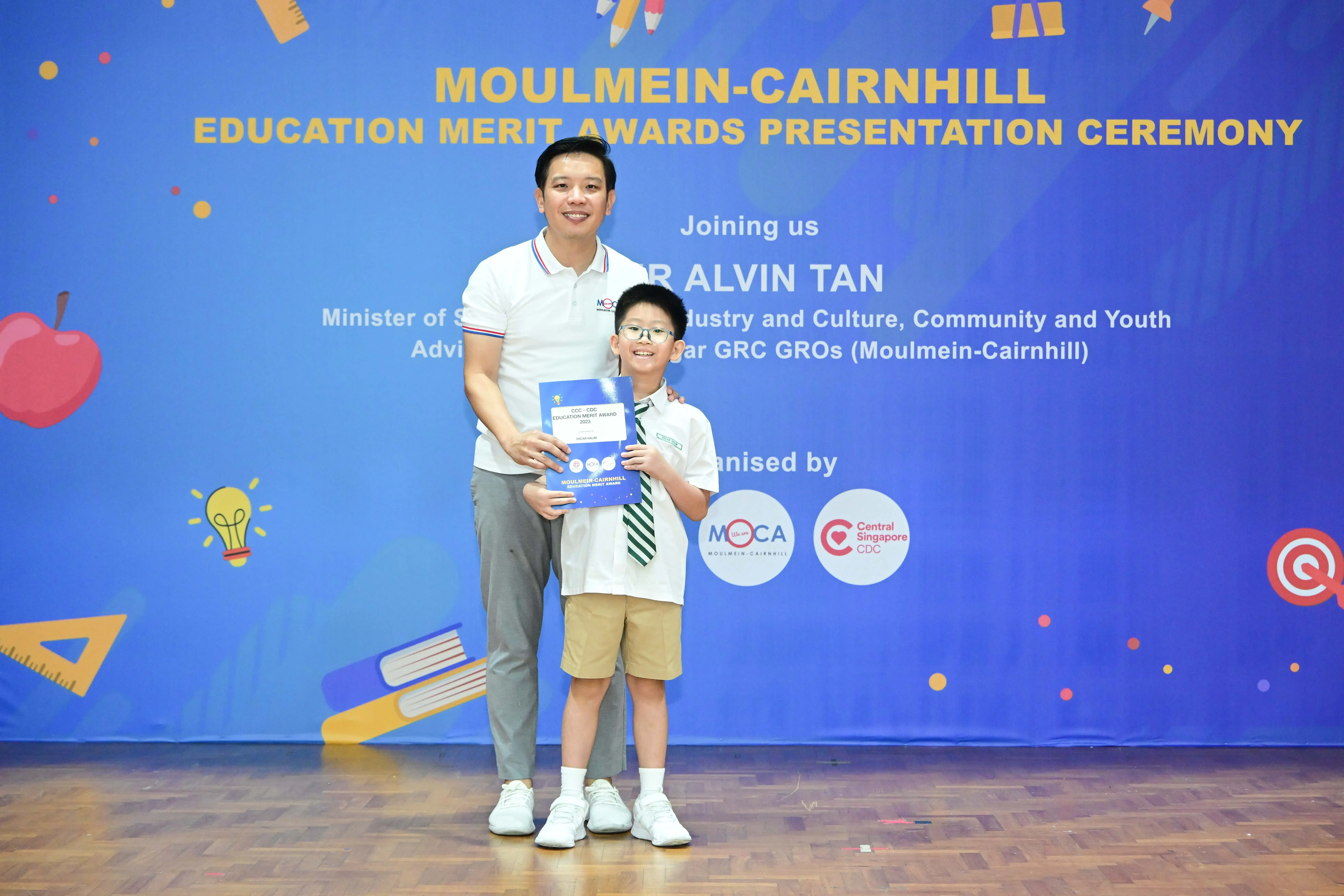 A man and a young boy holding an award certificate stand before a blue Moulmein-Cairnhill Education Merit Awards banner.