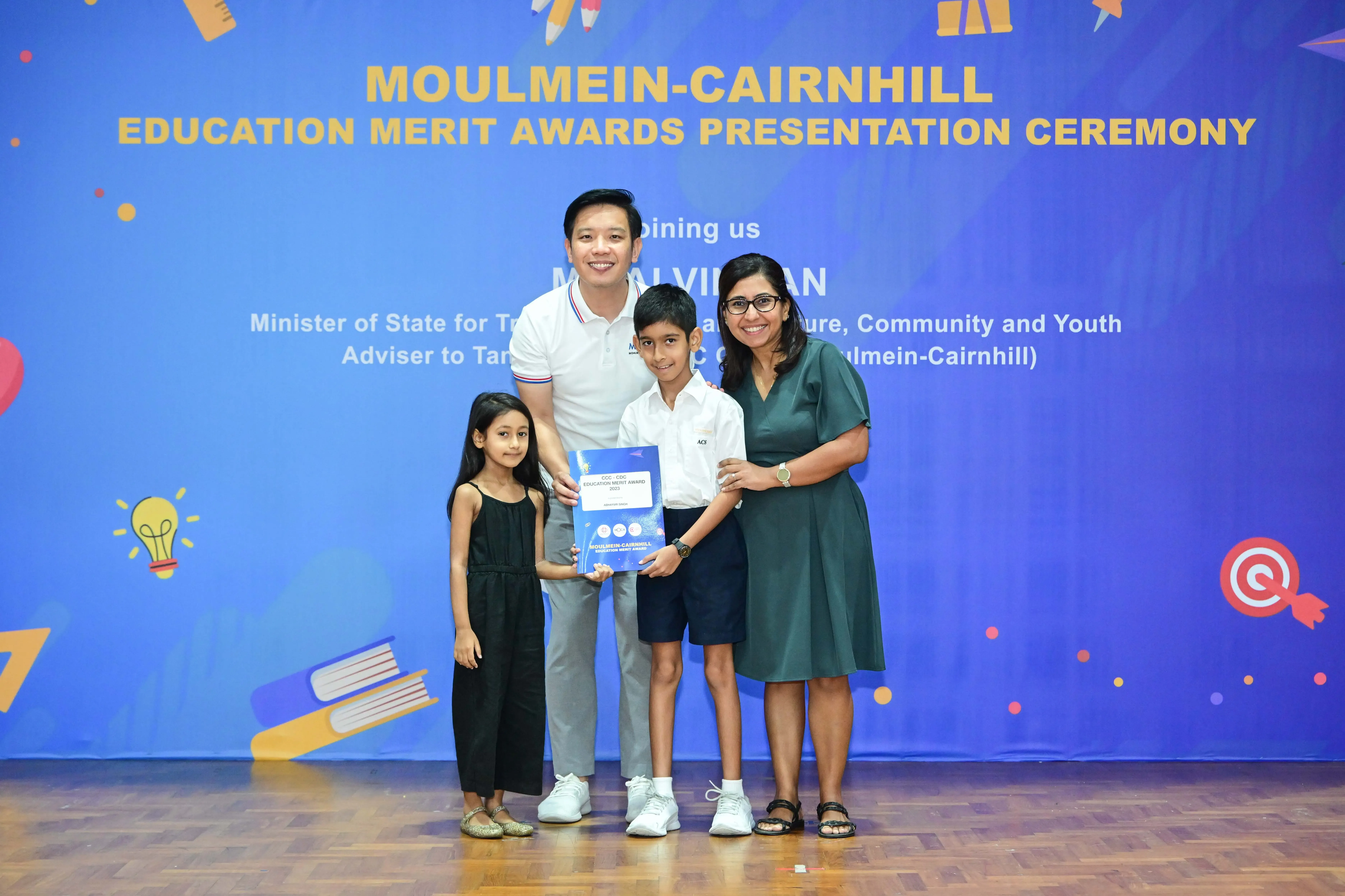 Four people pose with an award booklet in front of a Moulmein-Cairnhill Education Merit Awards banner.