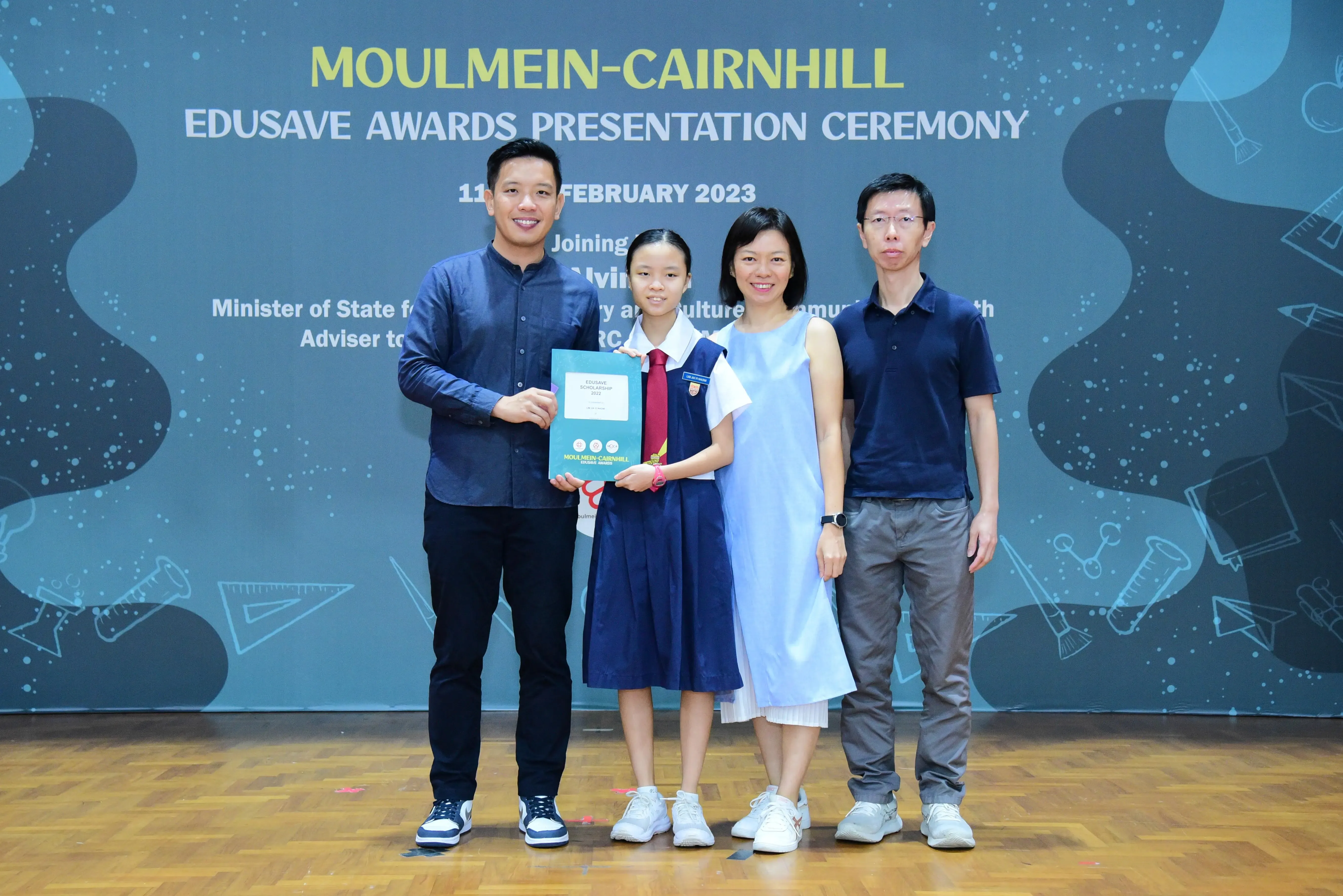 Four people pose at the Moulmein-Cairnhill Edusave Awards Ceremony; a girl in uniform holds a teal certificate folder.