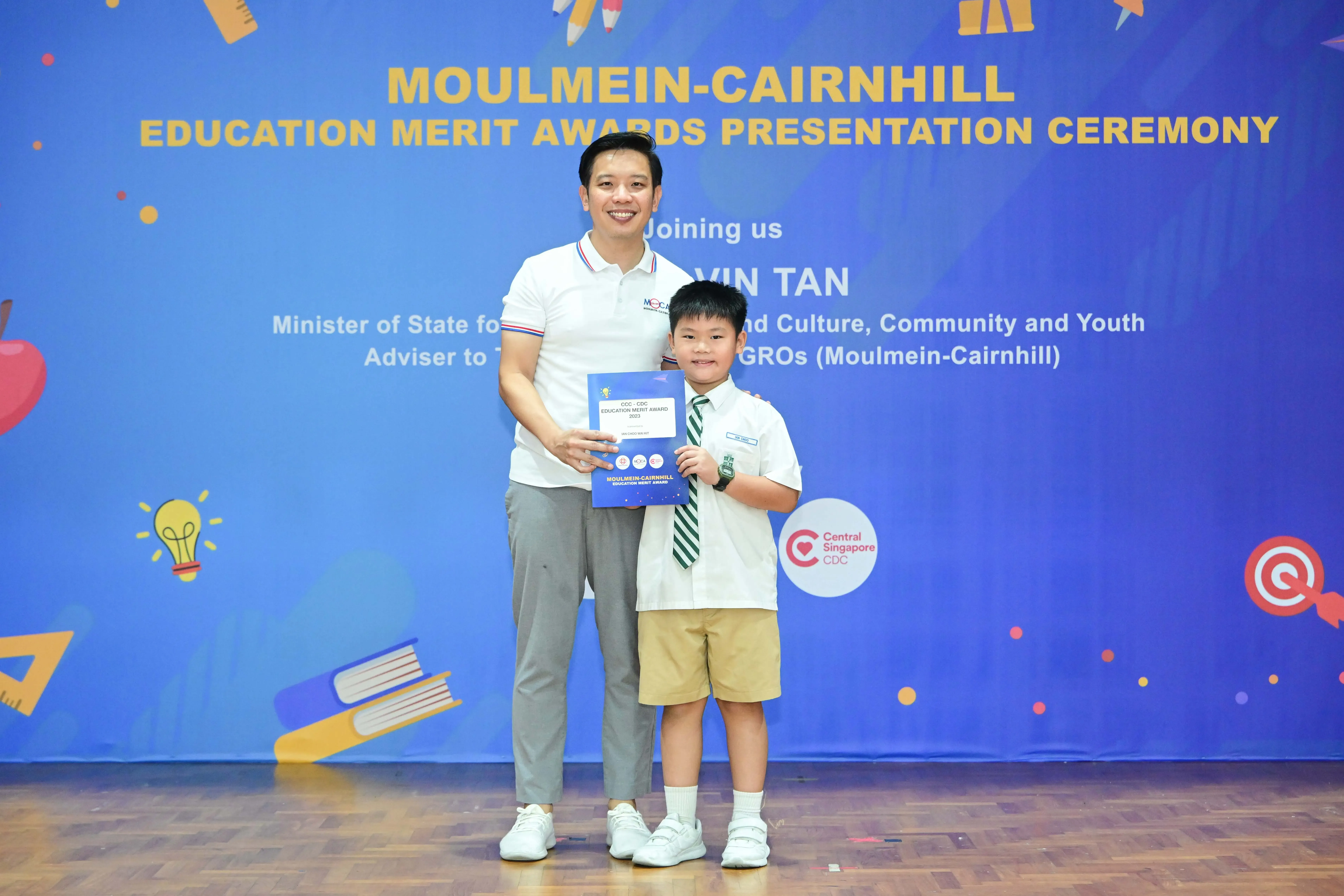 Man and young boy in school uniform hold an award certificate at Moulmein-Cairnhill Education Merit Awards ceremony.