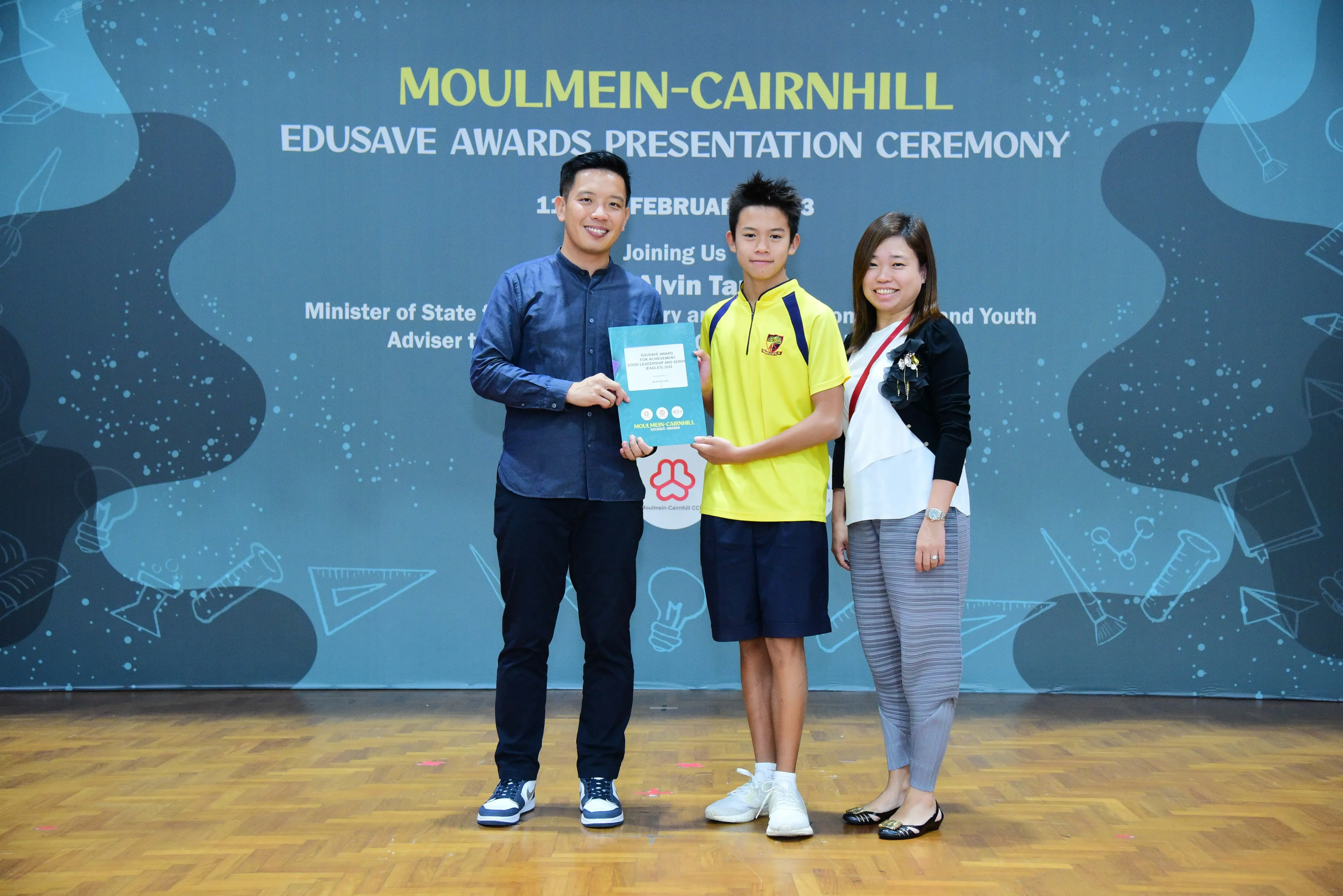 Three people stand smiling before a Moulmein-Cairnhill Edusave Awards Presentation Ceremony banner, holding a certificate.