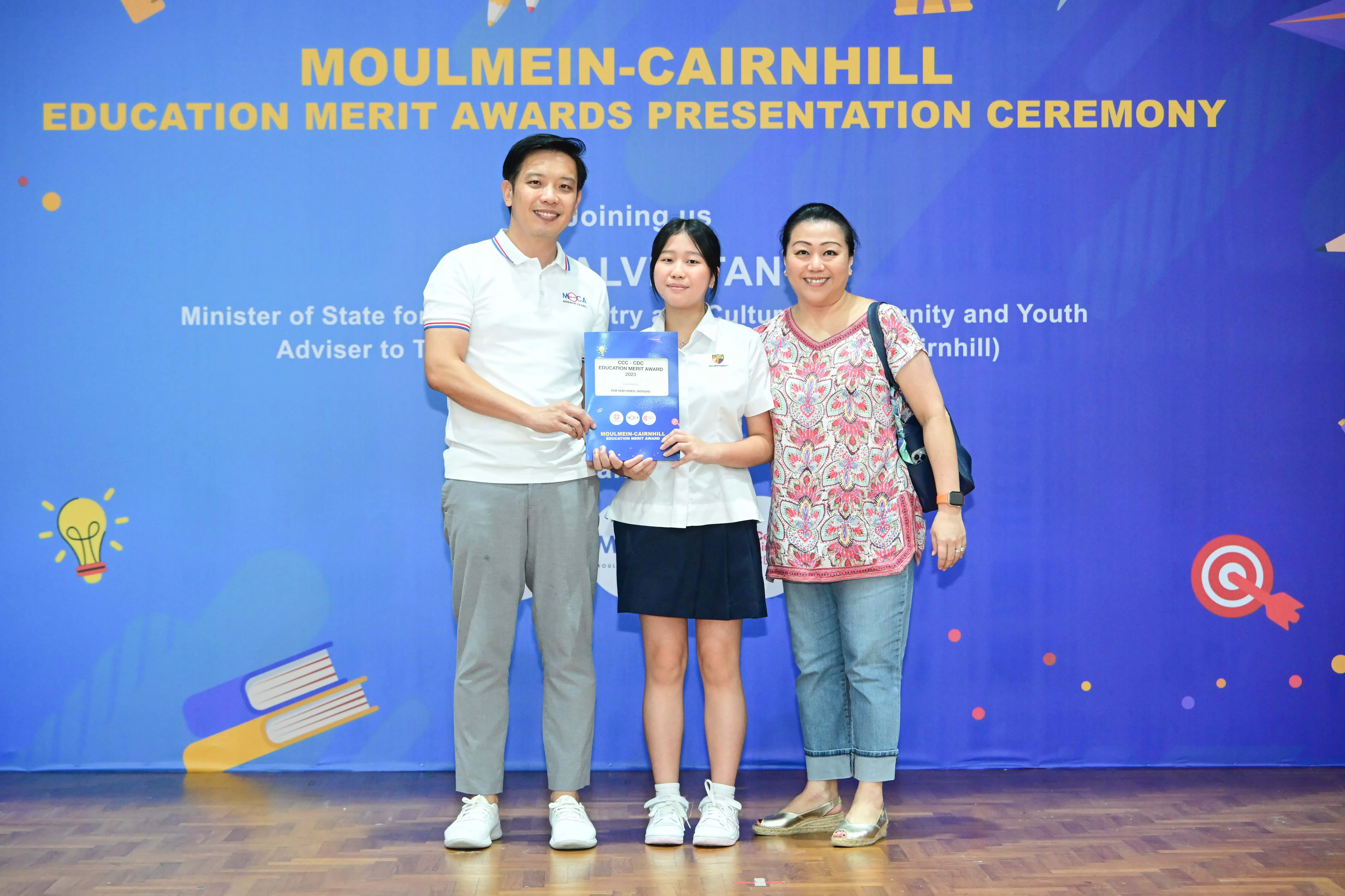 Three people pose holding an award certificate at the Moulmein-Cairnhill Education Merit Awards Presentation Ceremony.