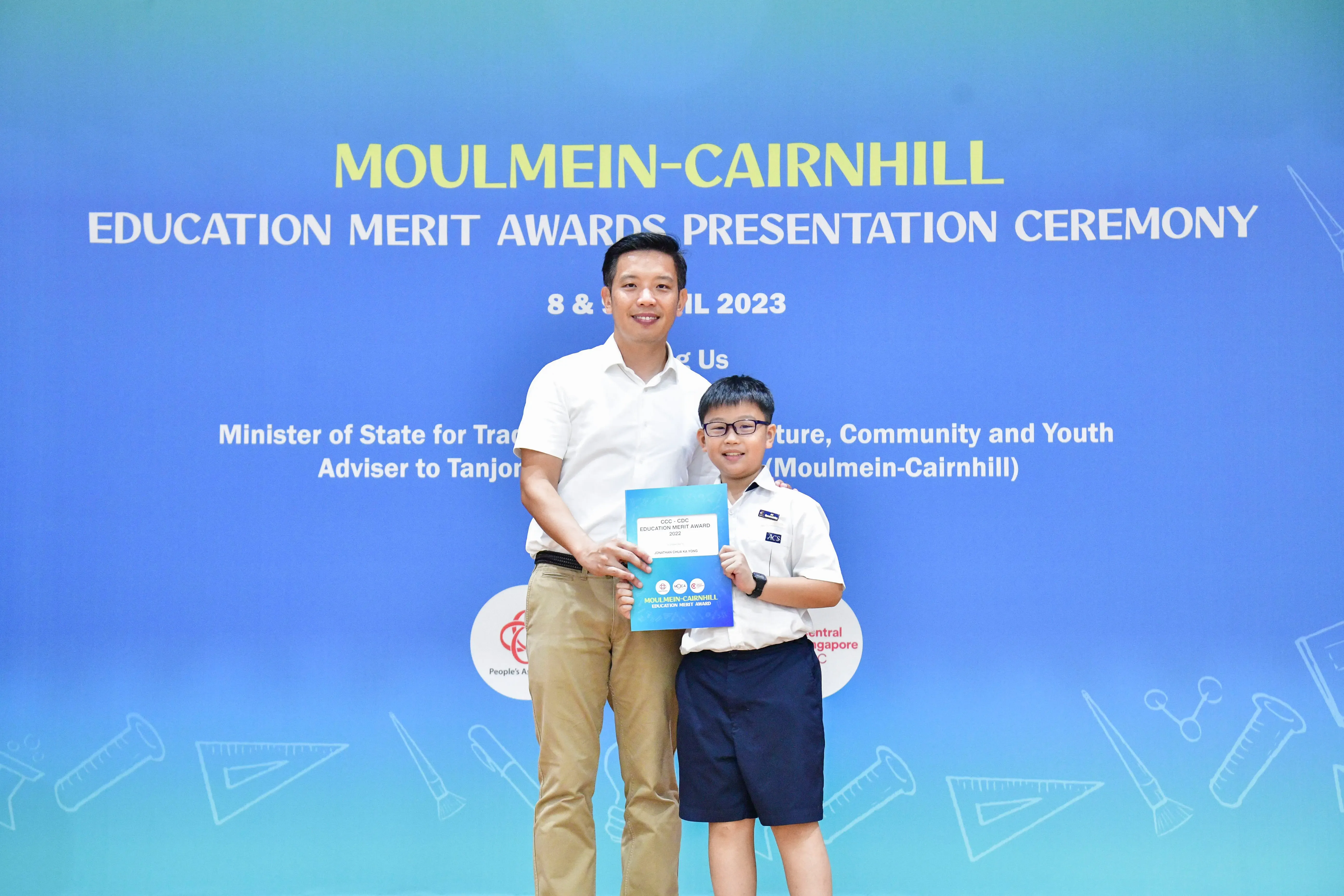 An adult and a young boy in school uniform hold an award certificate at the Moulmein-Cairnhill Education Merit Awards 2023.