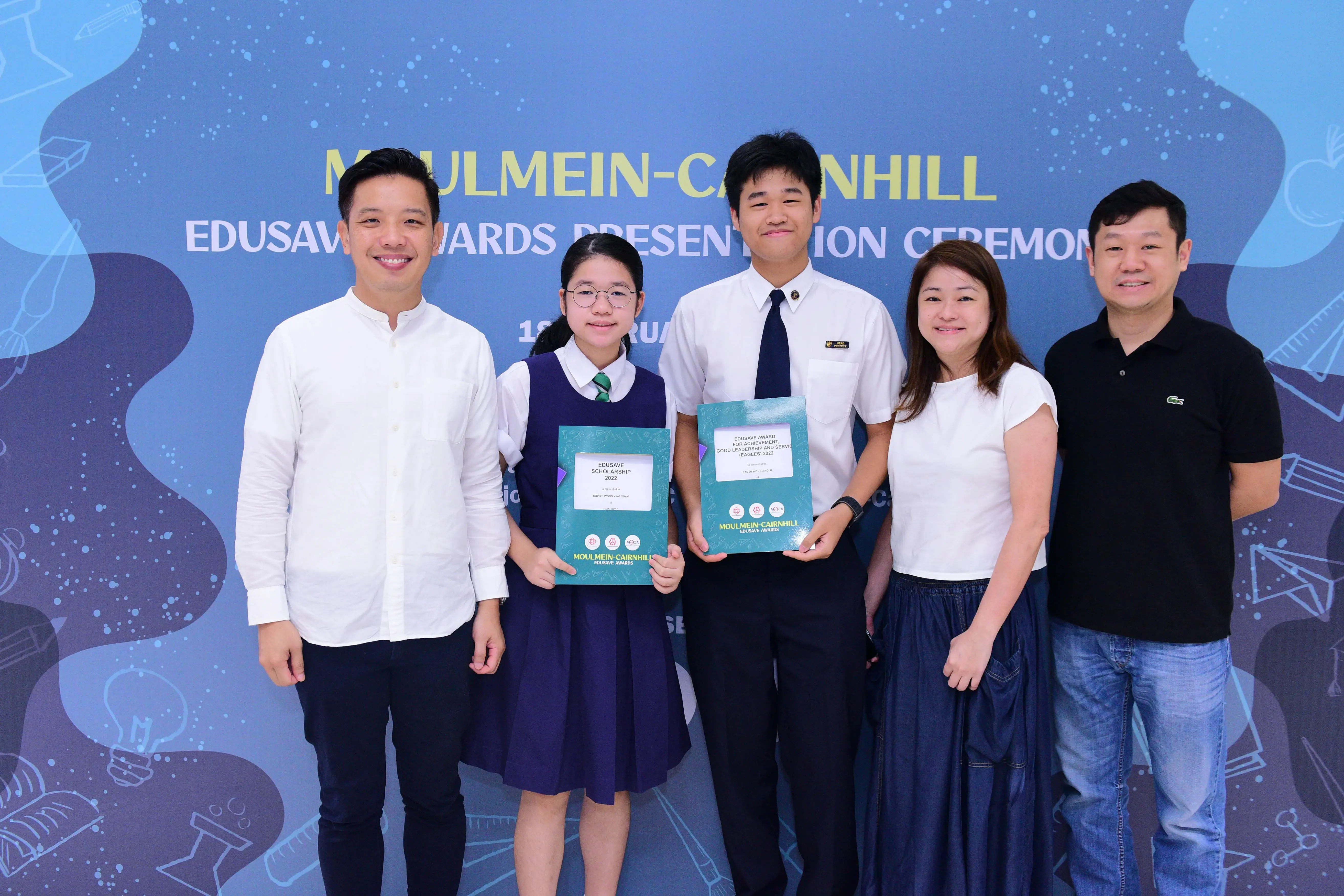 Five people pose smiling at the Moulmein-Cairnhill Edusave Awards Presentation Ceremony, two students holding award folders.