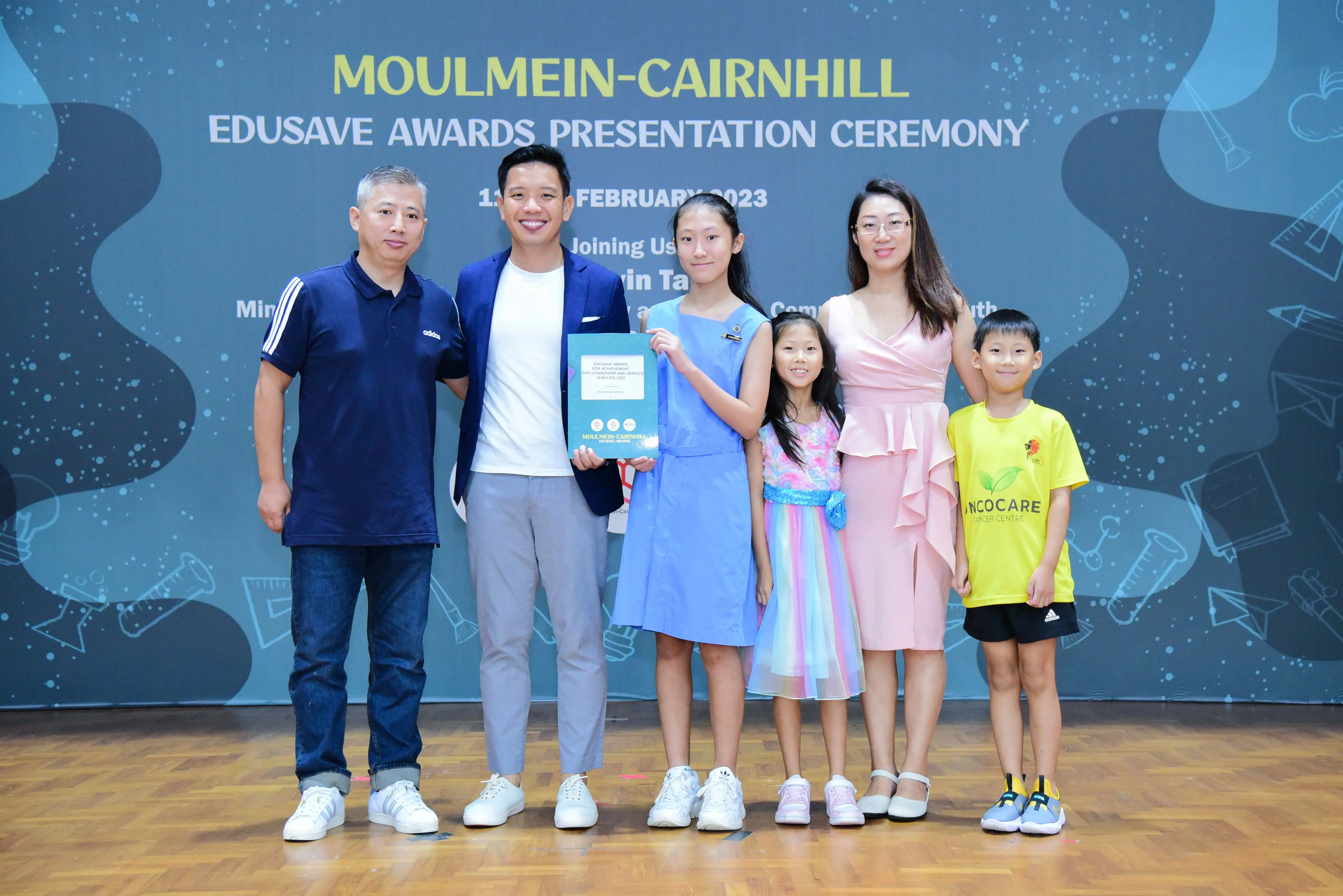 Six people pose at the Moulmein-Cairnhill Edusave Awards Presentation Ceremony in February 2023, with a girl holding a certificate.