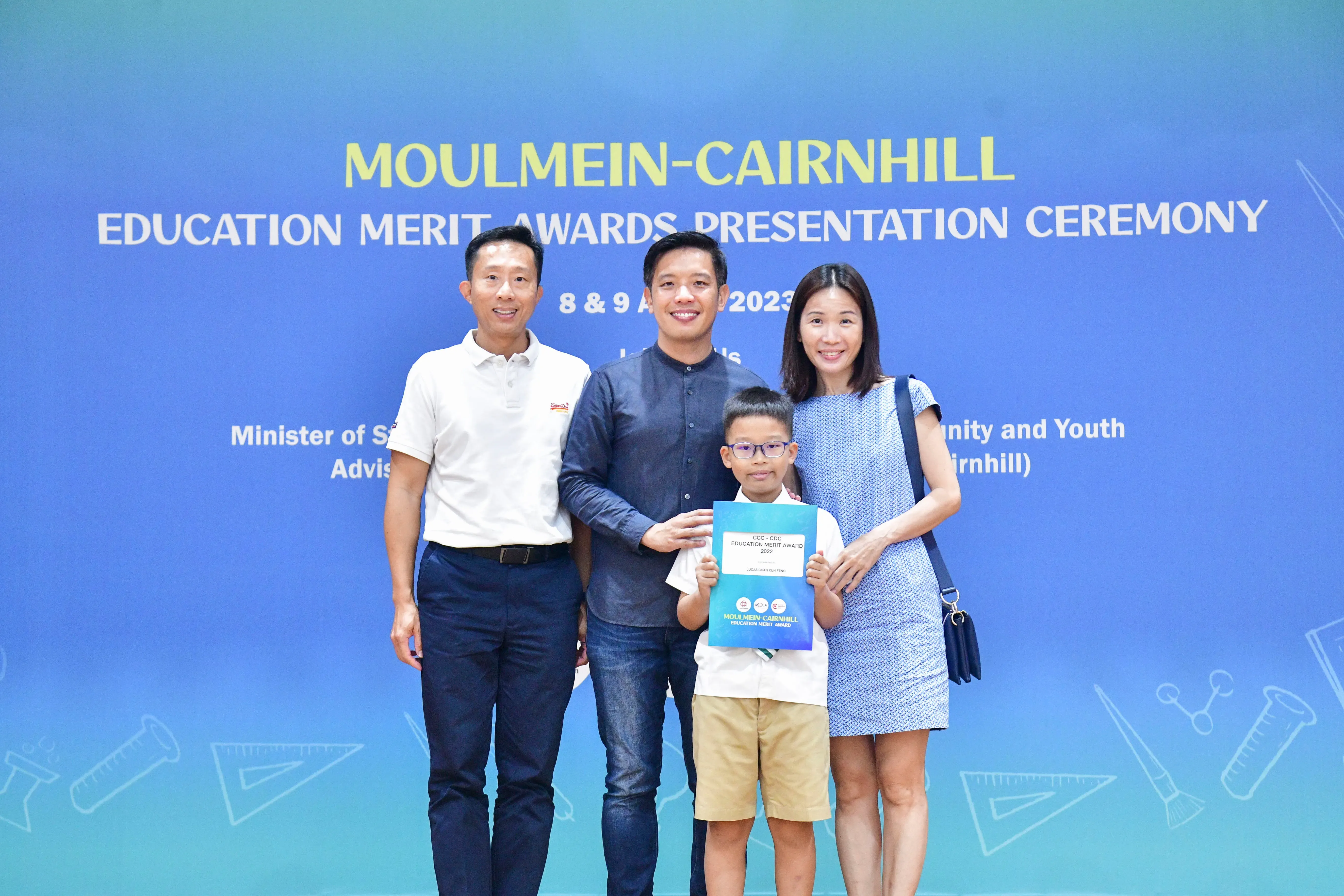 Three adults and a boy holding an award certificate pose before a Moulmein-Cairnhill Education Merit Awards backdrop.