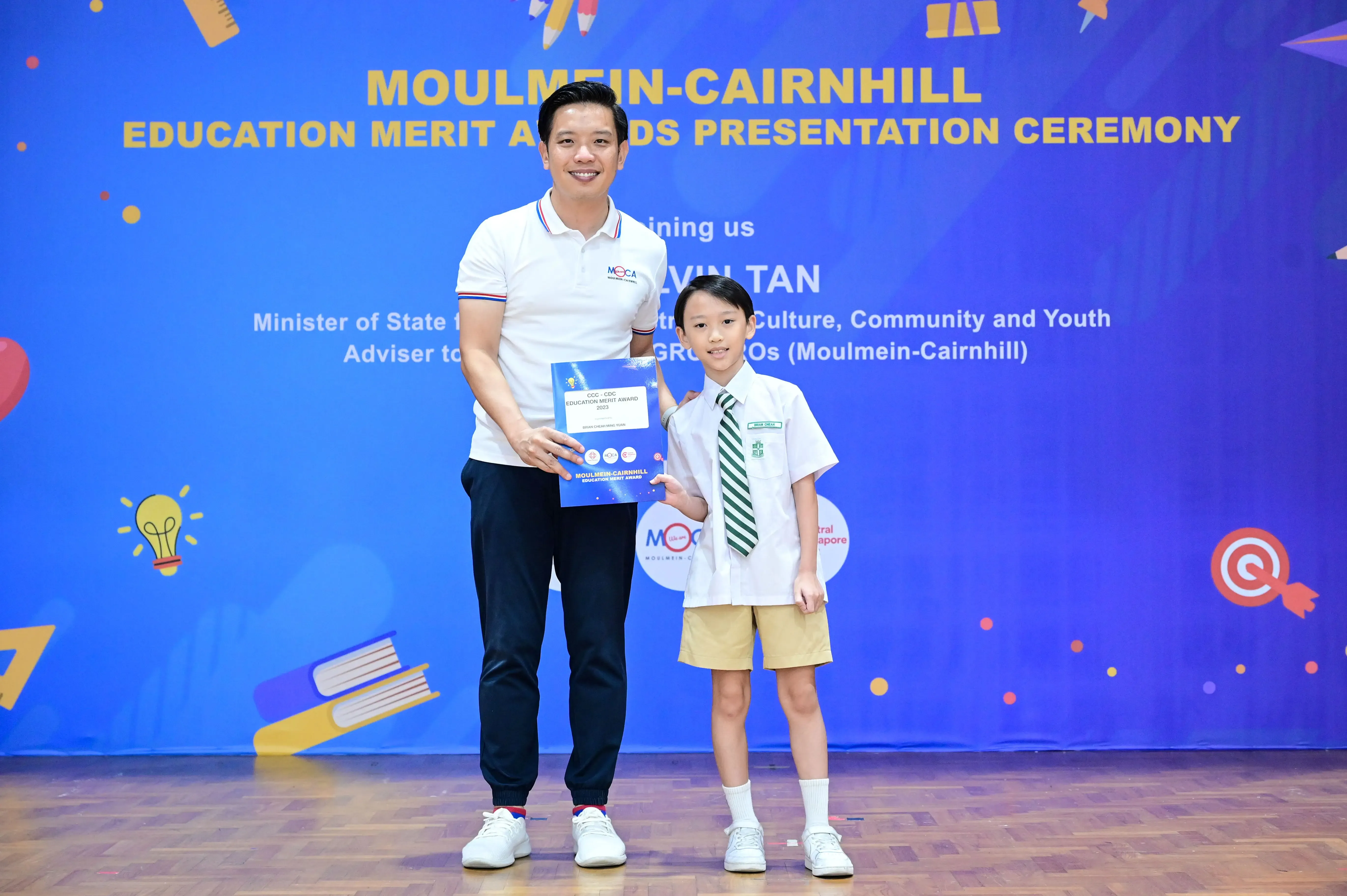 An adult and a young student in school uniform hold an award certificate at the Moulmein-Cairnhill Education Merit Awards ceremony.