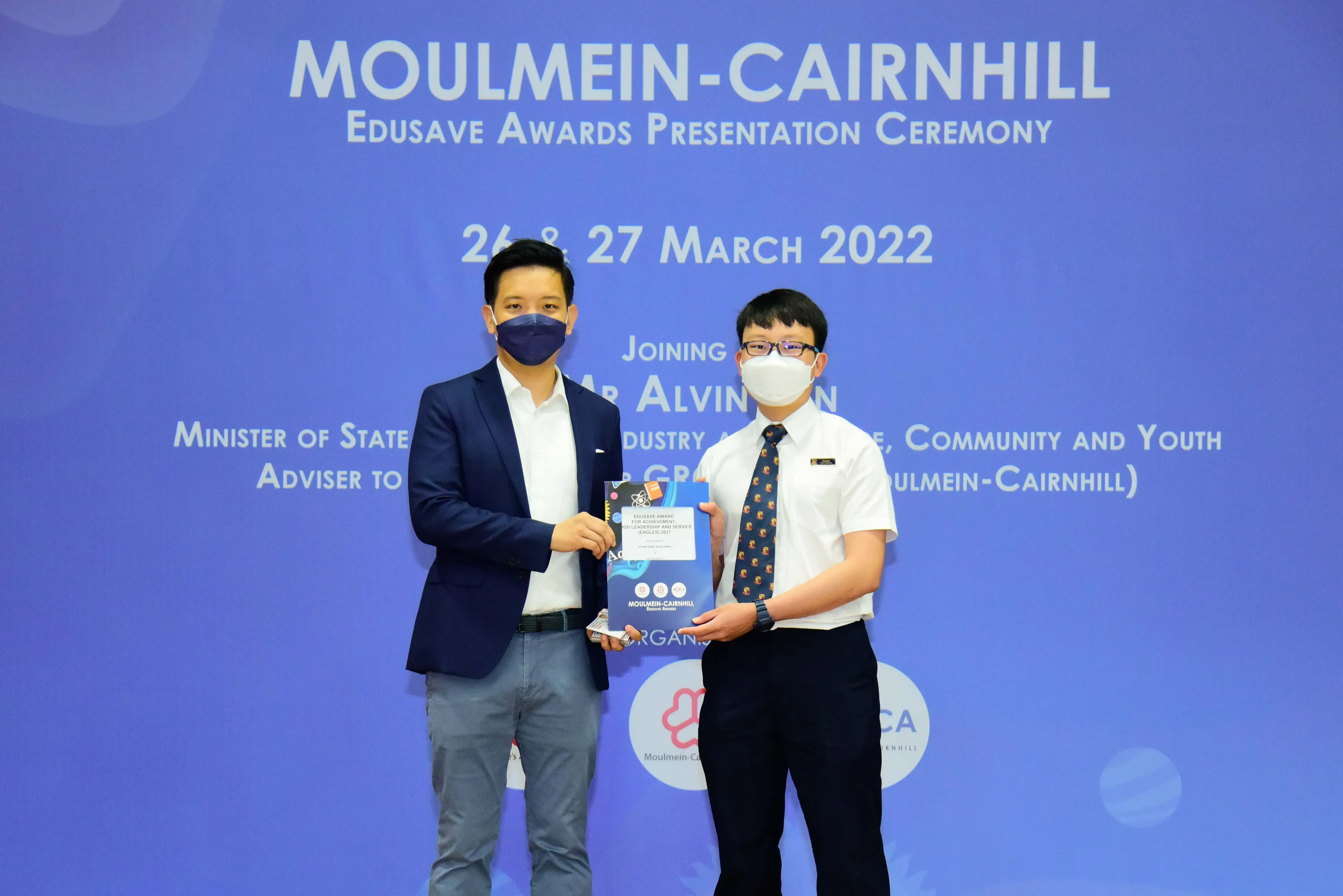 Two masked men hold an award booklet at the Moulmein-Cairnhill Edusave Awards Presentation Ceremony, March 2022.