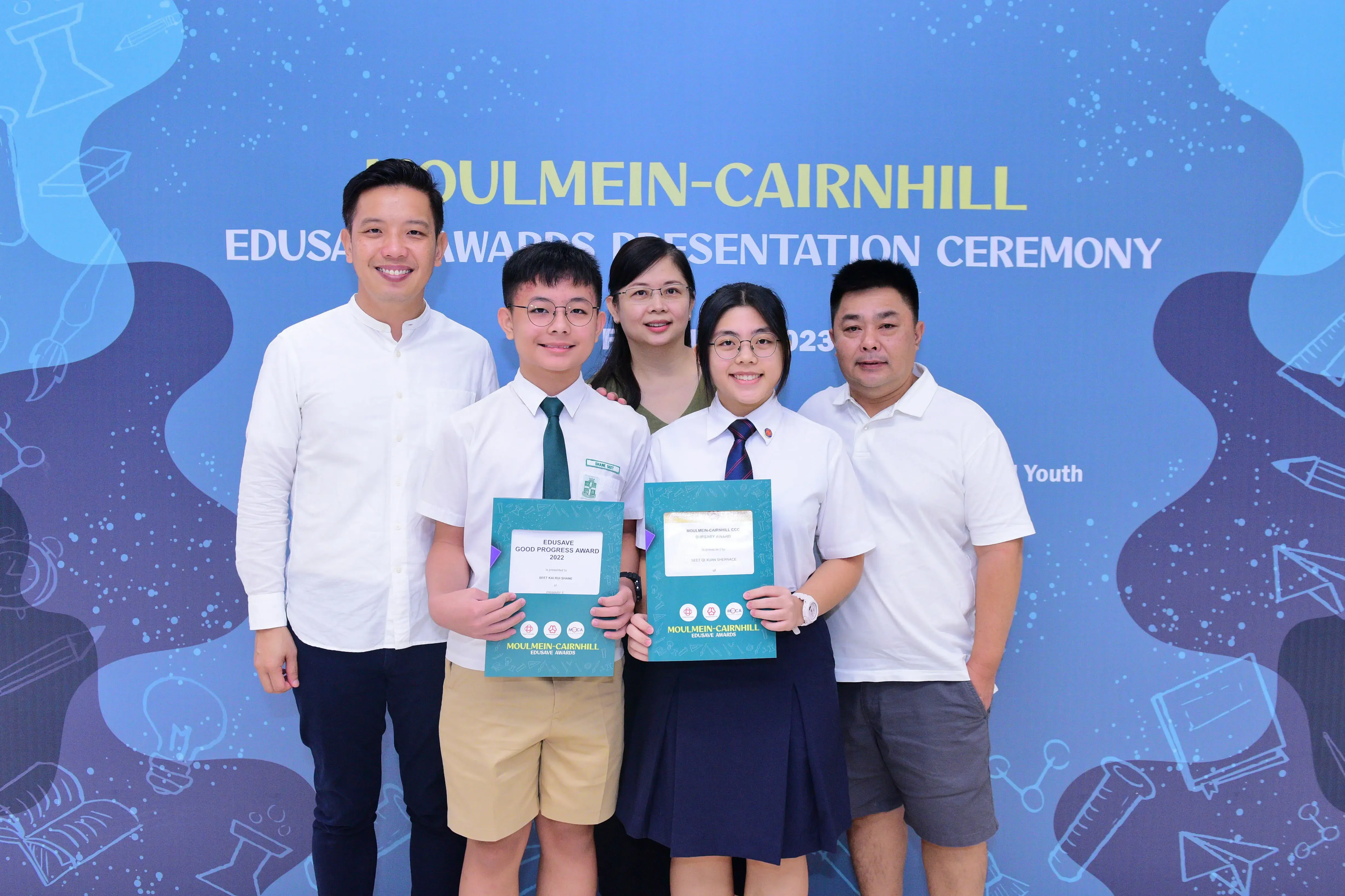Five people pose at the Moulmein-Cairnhill Edusave Awards Ceremony; two students hold teal award folders.