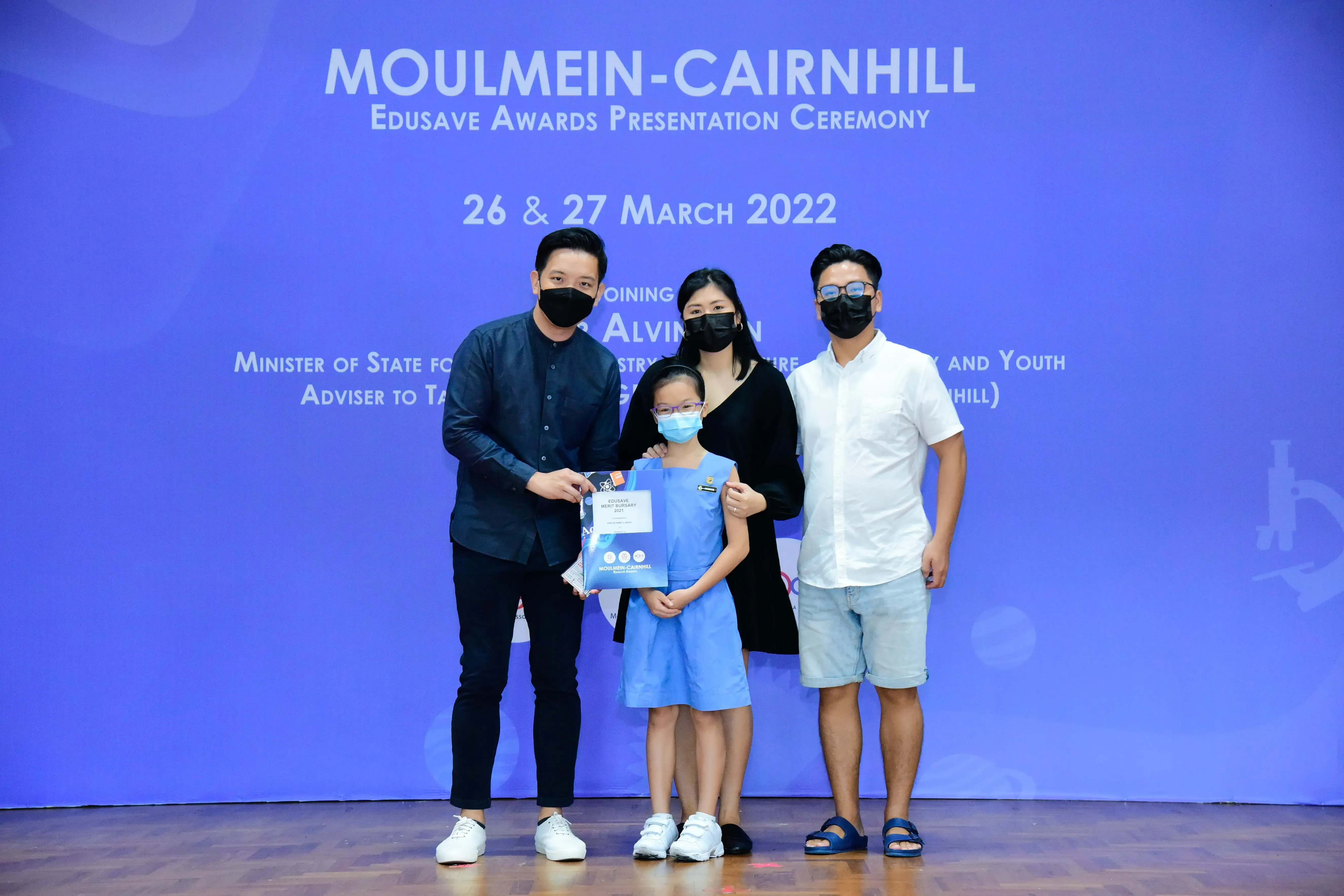 Three masked adults and a young girl holding an award at the Moulmein-Cairnhill Edusave Awards Ceremony, March 2022.