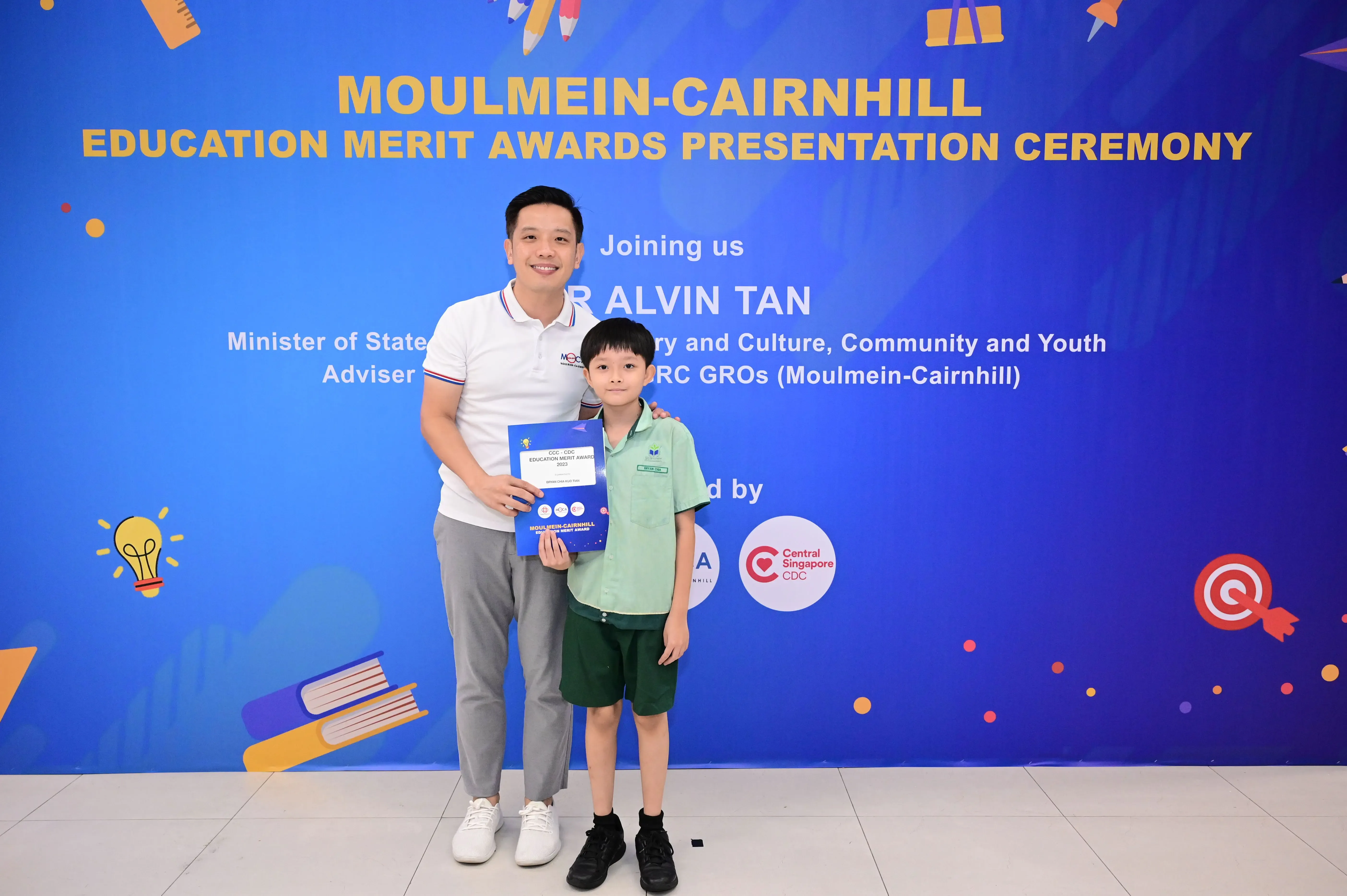 A man and a young boy pose holding an award in front of the Moulmein-Cairnhill Education Merit Awards banner.
