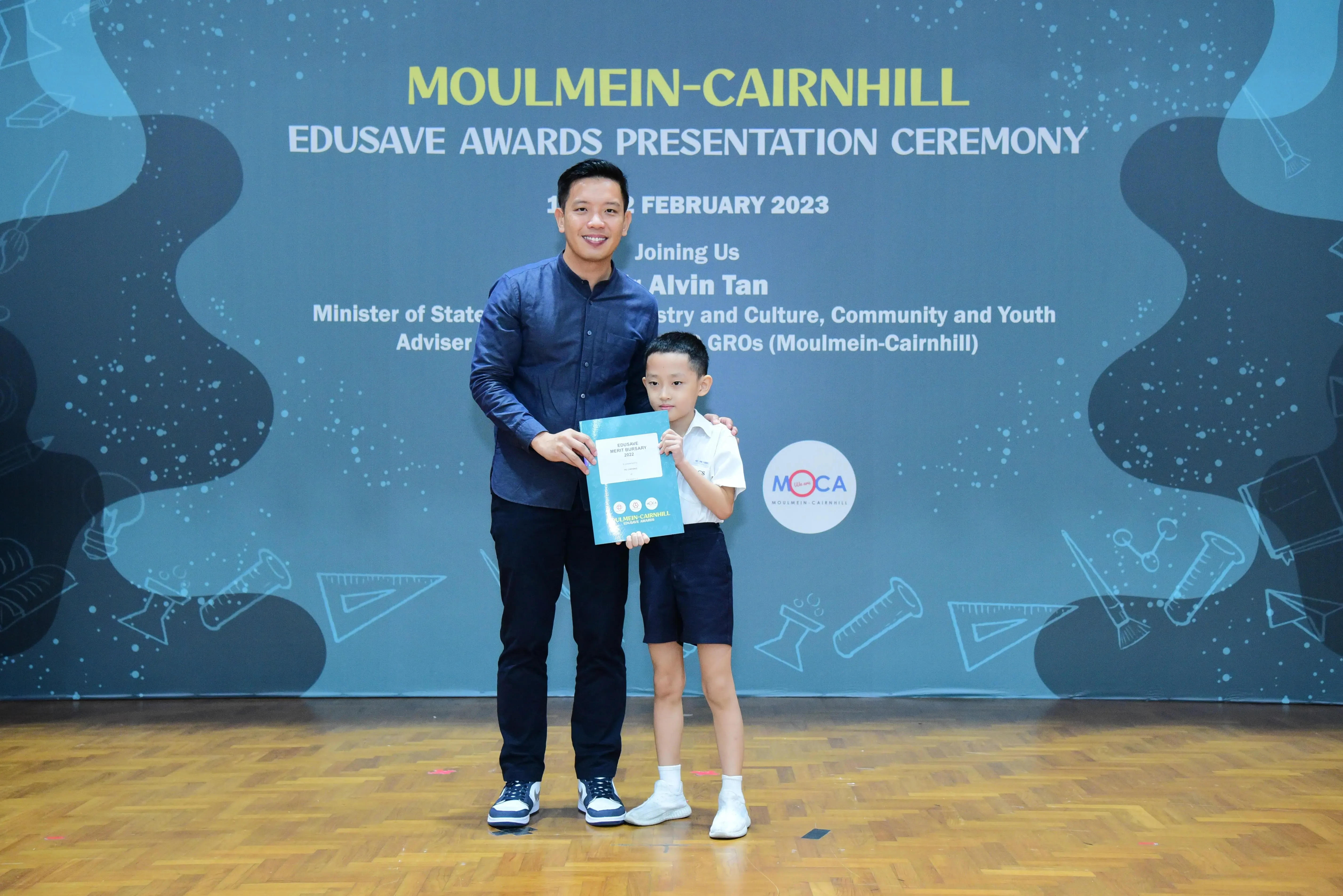 A man and a young boy hold a certificate together on stage at the Moulmein-Cairnhill Edusave Awards Ceremony, February 2023.