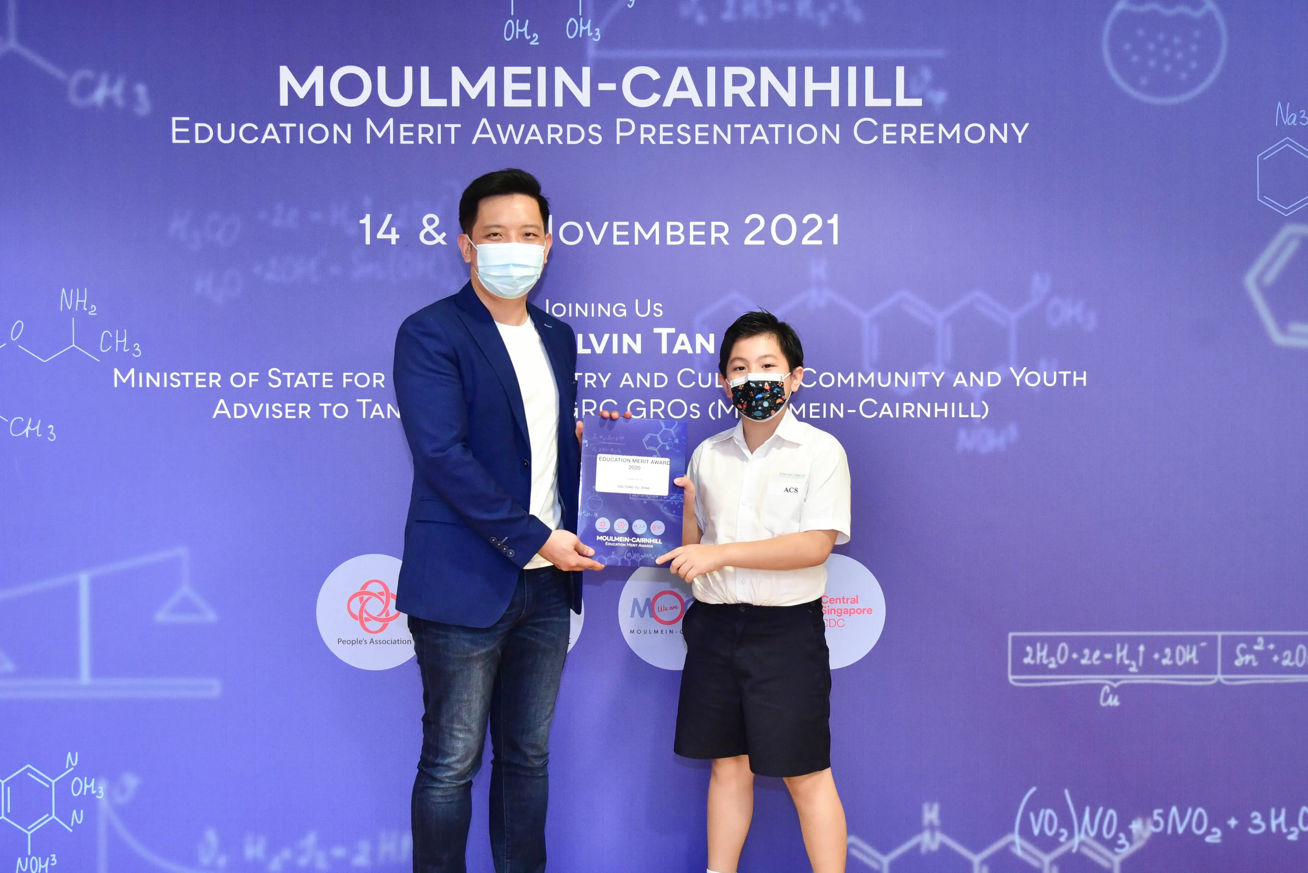 An adult and a child wearing masks hold an award certificate at the Moulmein-Cairnhill Education Merit Awards 2021.