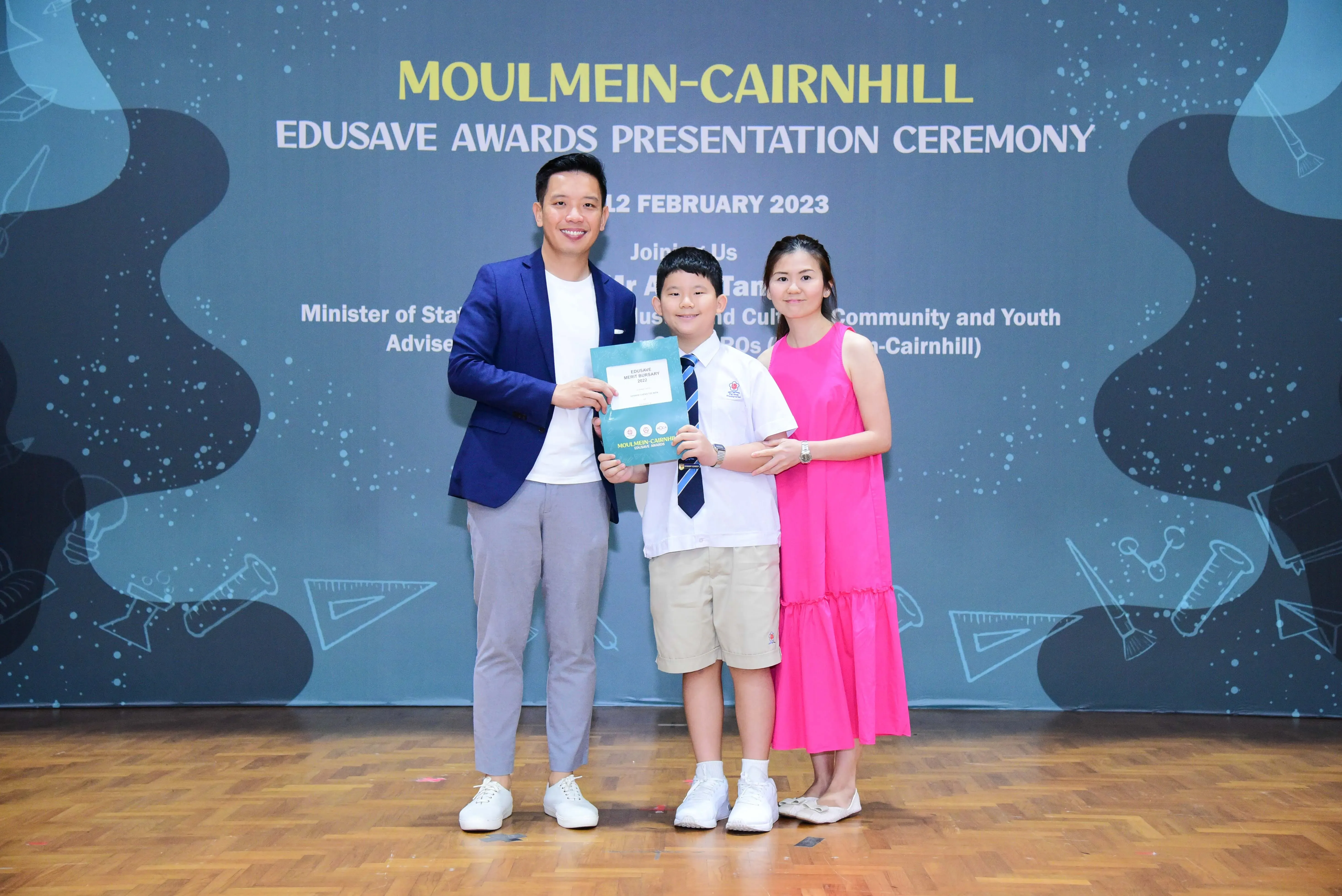 A boy in school uniform holds a certificate between two adults at the Moulmein-Cairnhill Edusave Awards Ceremony 2023.