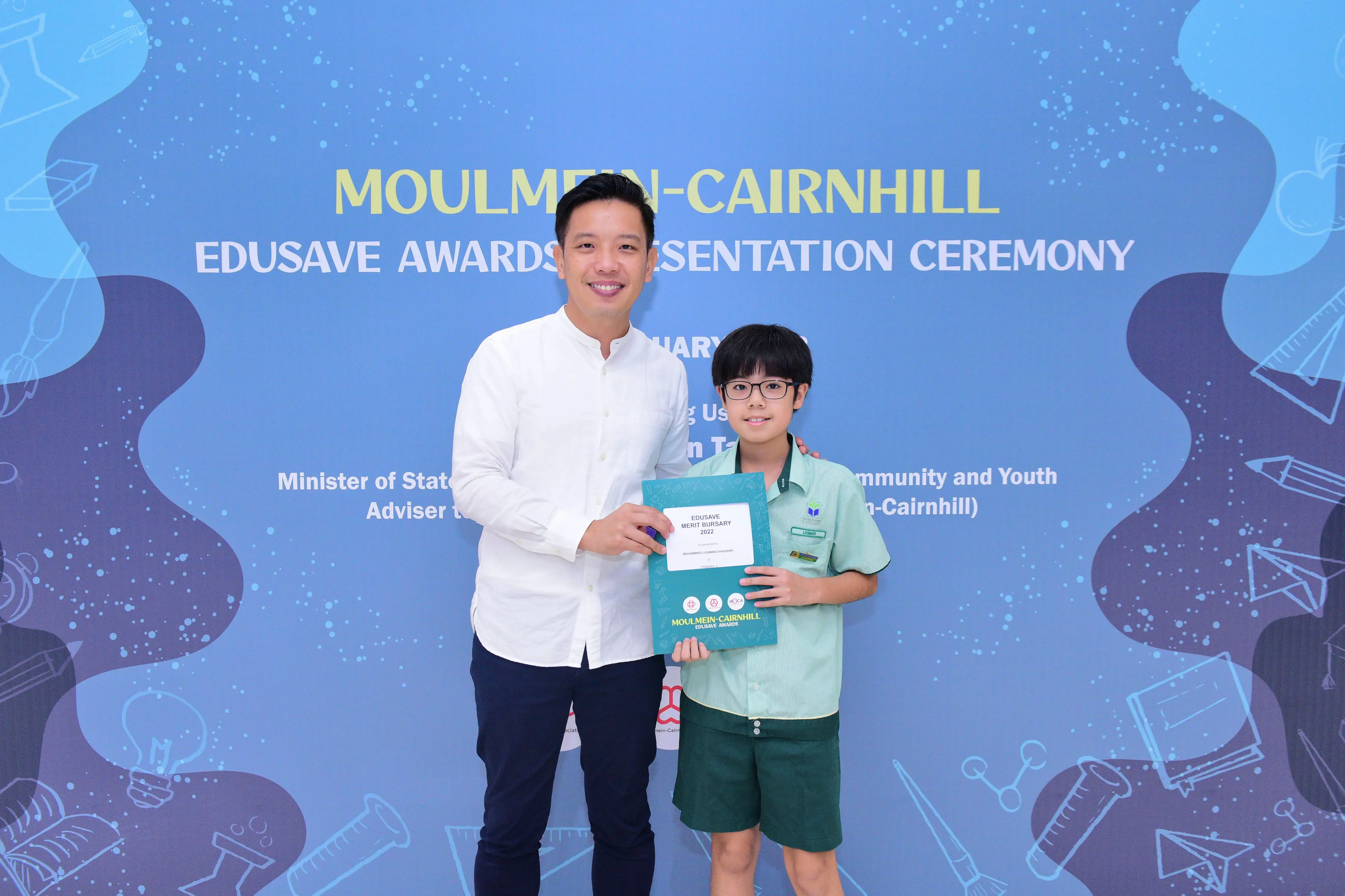 A man and a young boy in school uniform hold an Edusave Merit Bursary certificate at the Moulmein-Cairnhill awards ceremony.