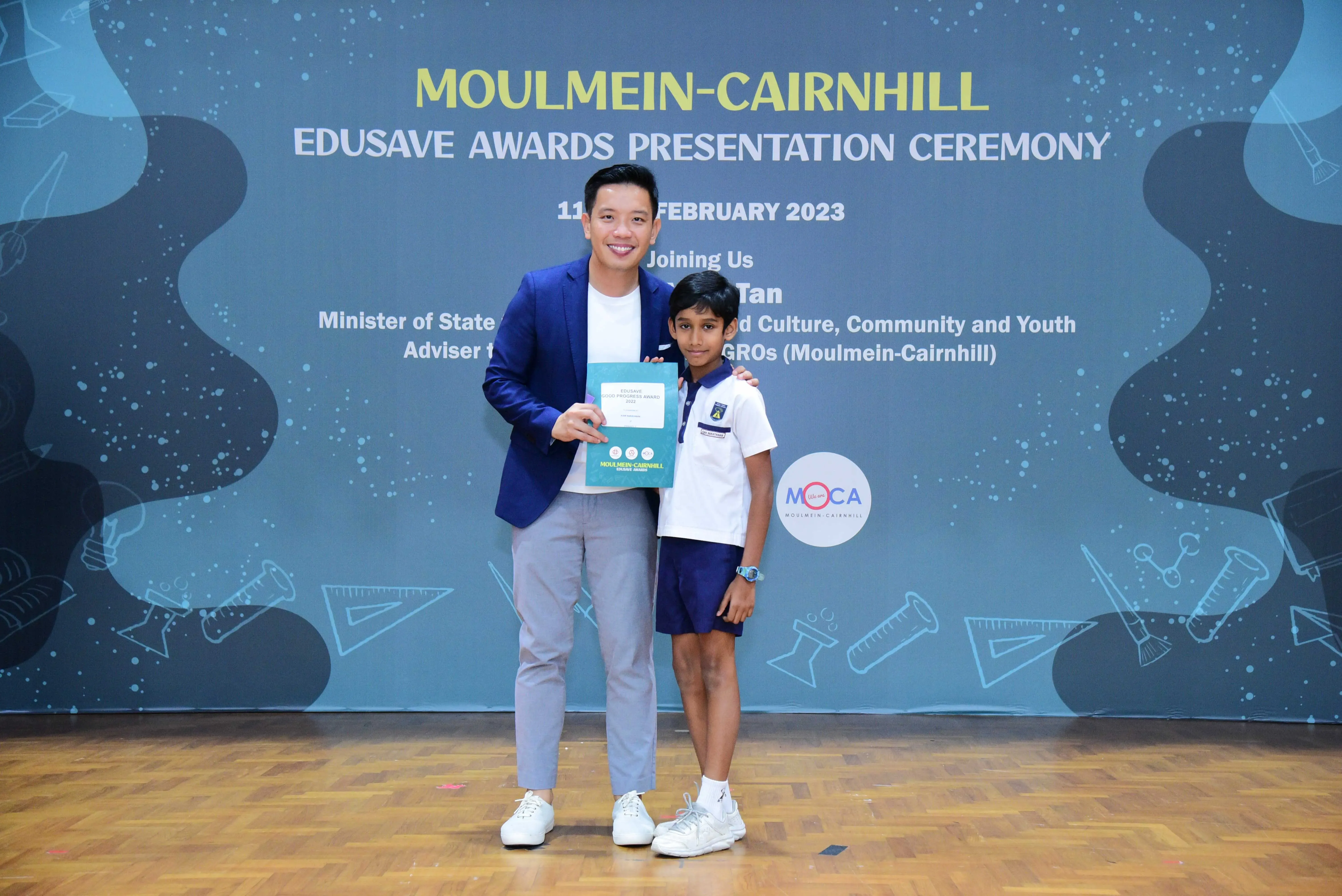 Adult in a blue blazer poses with a young student holding an award at the Moulmein-Cairnhill Edusave Awards Ceremony 2023.